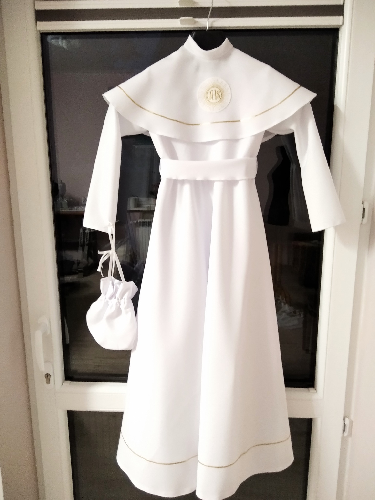 Biała alba komunijna z kapturem i torebką, zawieszona na drzwiach, wykończona złotą nicią.