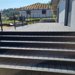 Schody z ciemnych desek kompozytowych prowadzące na taras, w tle budynek w trakcie budowy i zieleń