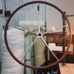 Drewniana kierownica jachtowa z metalowymi szprychami, w tle materiały tapicerskie i narzędzia w warsztacie.