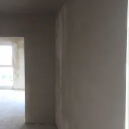 Świeżo nałożona, jasnoszara warstwa tynku na ścianach wewnętrznych pomieszczenia w stanie surowym, widoczne puszki elektryczne, zarys okna w tle.