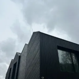 Nowoczesna elewacja domu z ciemnego drewna, widok z dołu na tle pochmurnego nieba, z widocznymi oknami i elementami konstrukcyjnymi.