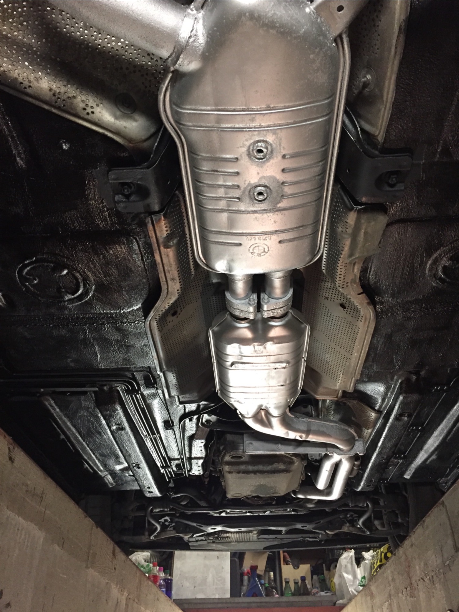 Spód samochodu po zabezpieczeniu antykorozyjnym, widoczny układ wydechowy, elementy podwozia pokryte czarną powłoką ochronną, metalowe osłony termiczne, ujęcie od dołu z kanału warsztatowego.