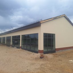 ELEWACJE Paweł Pietrzak - Budynek po tynkowaniu elewacji, z widocznymi oknami i fragmentami ceglanej podmurówki. Teren wokół budynku w trakcie prac wykończeniowych.