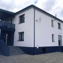 Dwukondygnacyjny dom z białą elewacją i ciemnoszarą podmurówką, balkonem i schodami. Widoczne przewody na brukowanej powierzchni.