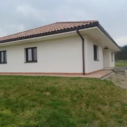 Jasny, parterowy dom z brązową dachówką, świeżo otynkowaną białą elewacją i brązowymi rynnami, otoczony trawnikiem i polną drogą.