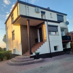 Dwukondygnacyjny dom z jasną elewacją, brązowymi elementami wykończeniowymi, schodami z kostki brukowej i widocznym klimatyzatorem na ścianie.