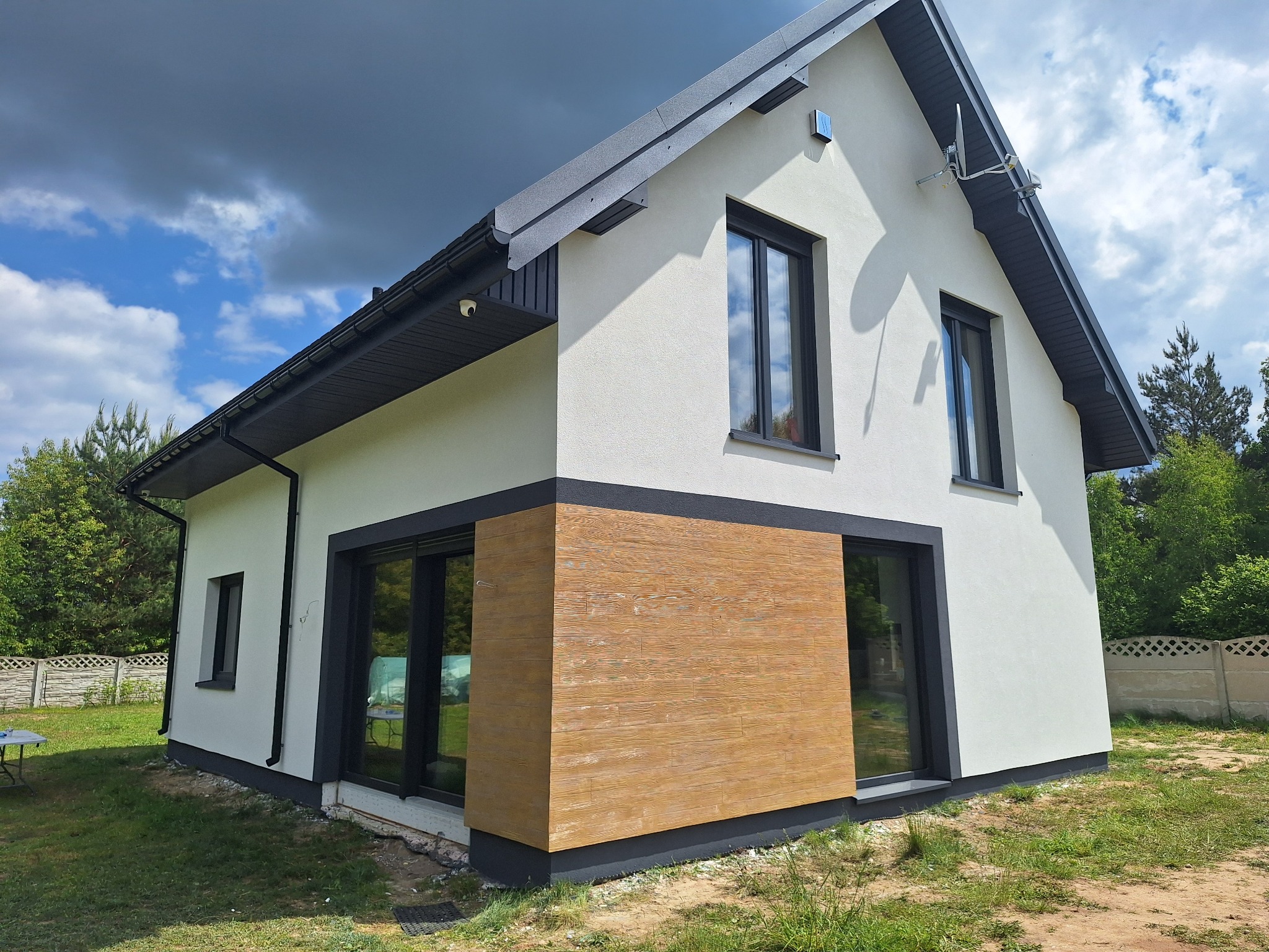 Nowoczesny dom z elewacją w kolorze złamanej bieli i drewnianą okładziną, czarne ramy okienne, widok z boku, trawnik i częściowo widoczne ogrodzenie.