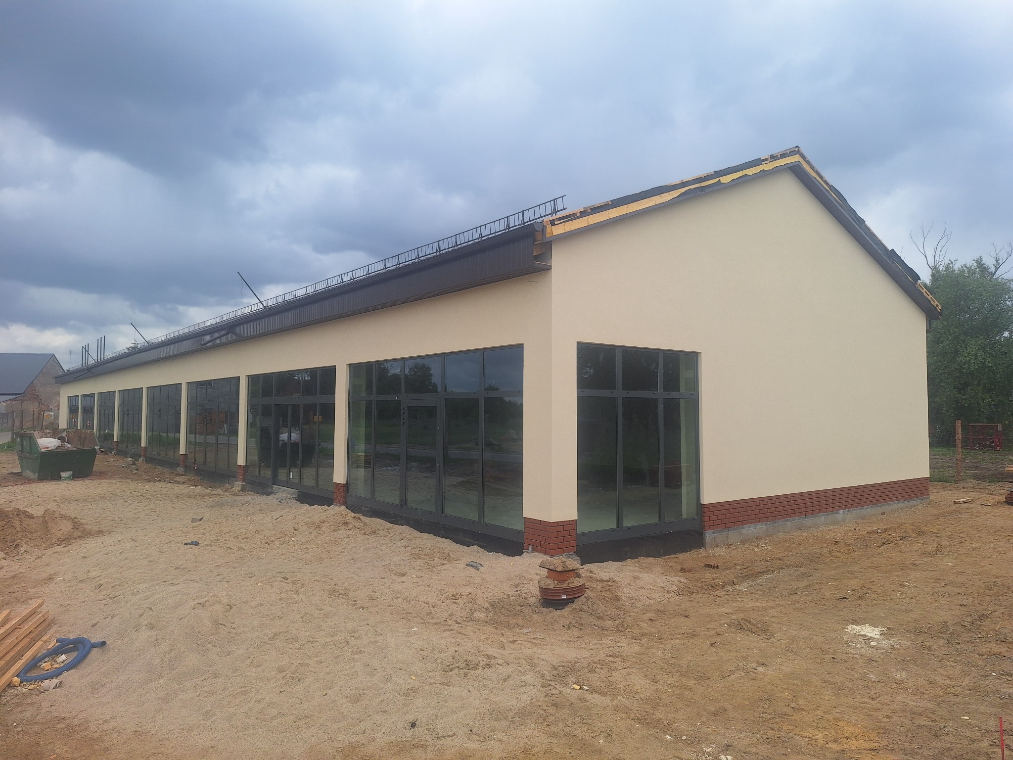 Budynek po tynkowaniu elewacji, z widocznymi oknami i fragmentami ceglanej podmurówki. Teren wokół budynku w trakcie prac wykończeniowych.