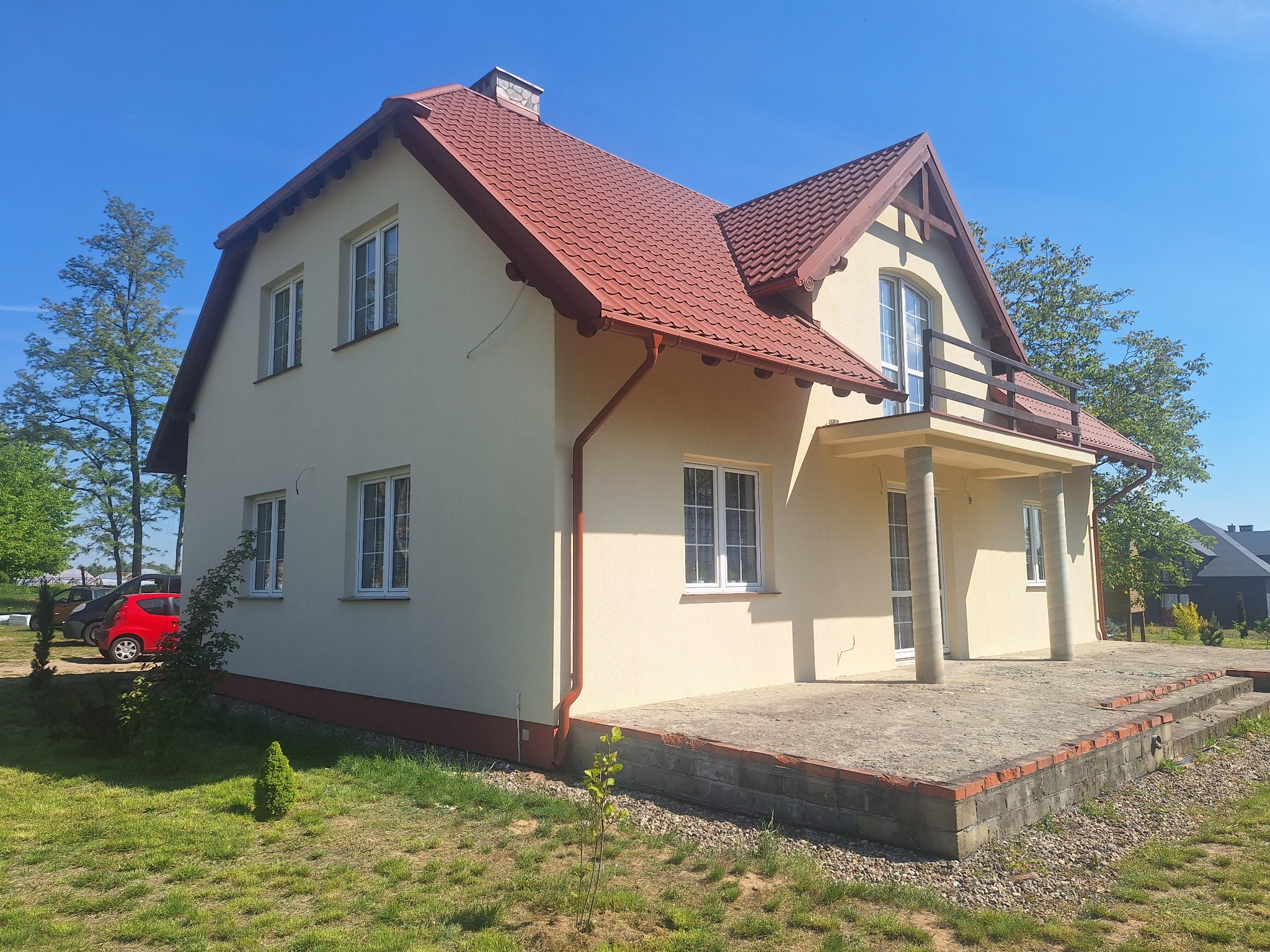 Jasny, dwukondygnacyjny dom z czerwonym dachem i balkonem, otoczony zielonym trawnikiem, w słoneczny dzień. Widoczny fragment czerwonego auta.