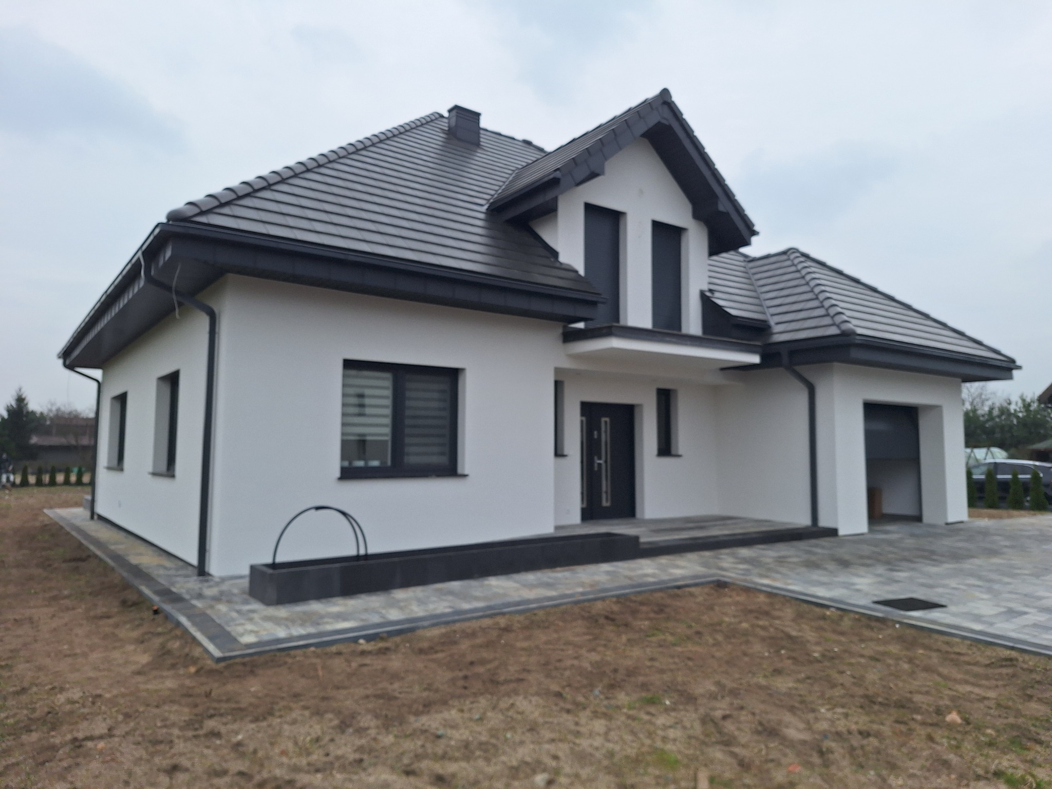 Nowoczesny dom jednorodzinny z białą elewacją i ciemnoszarą dachówką, widok z przodu. Minimalistyczna architektura, kostka brukowa na podjeździe.
