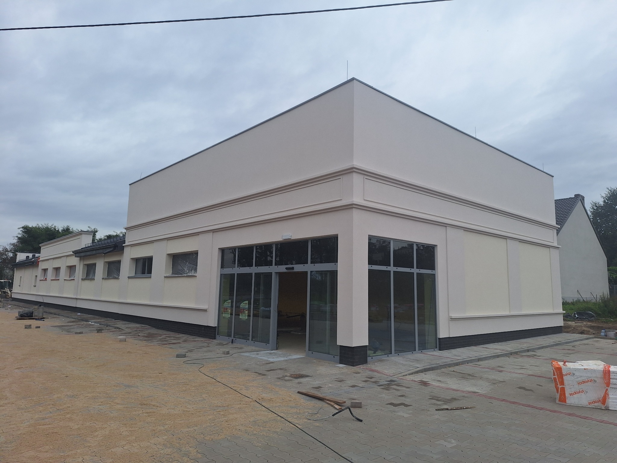 Jasna elewacja budynku komercyjnego z dużymi oknami i otwartymi drzwiami wejściowymi. Widoczne detale architektoniczne i kostka brukowa na zewnątrz.