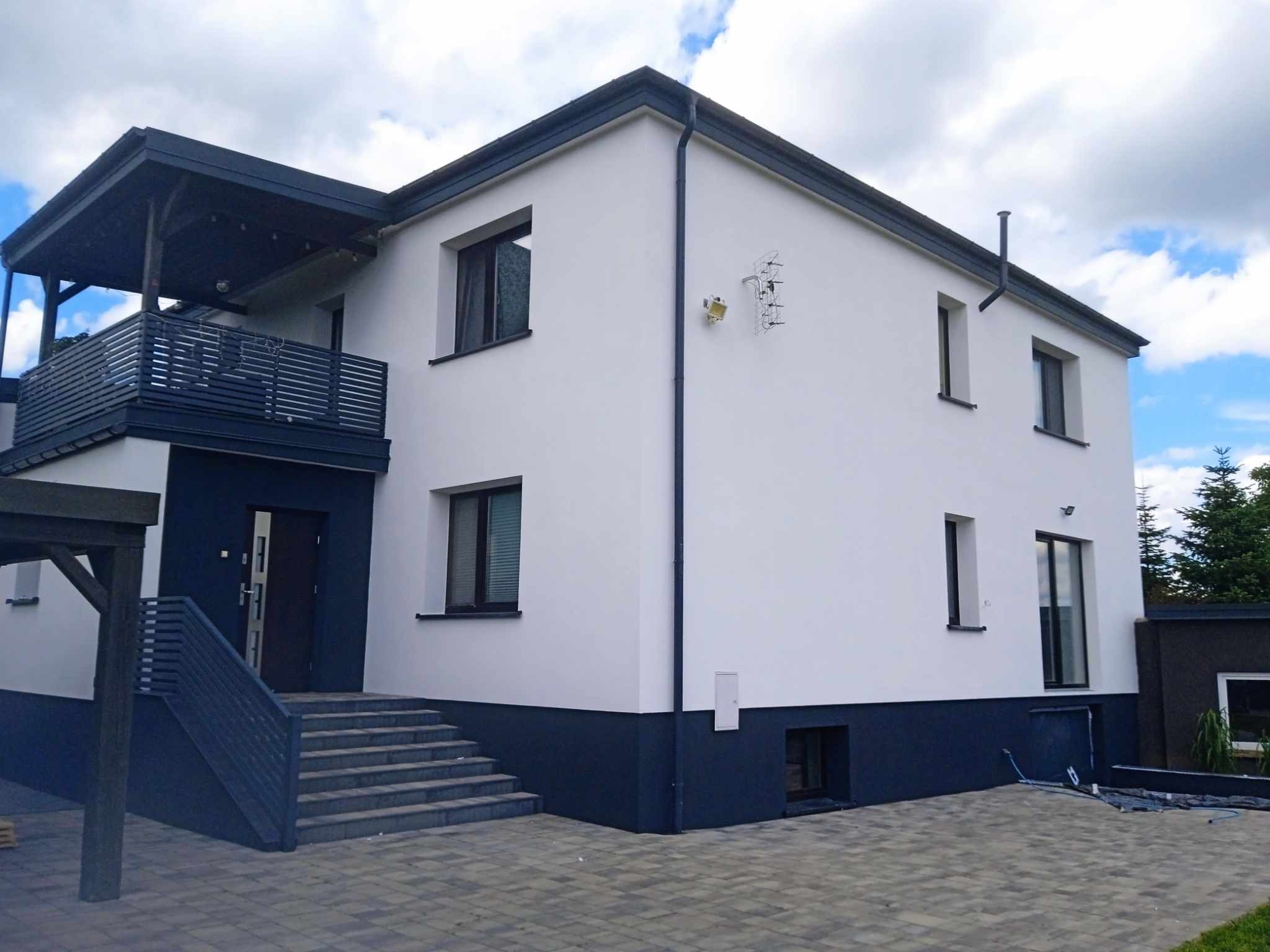 Dwukondygnacyjny dom z białą elewacją i ciemnoszarą podmurówką, balkonem i schodami. Widoczne przewody na brukowanej powierzchni.