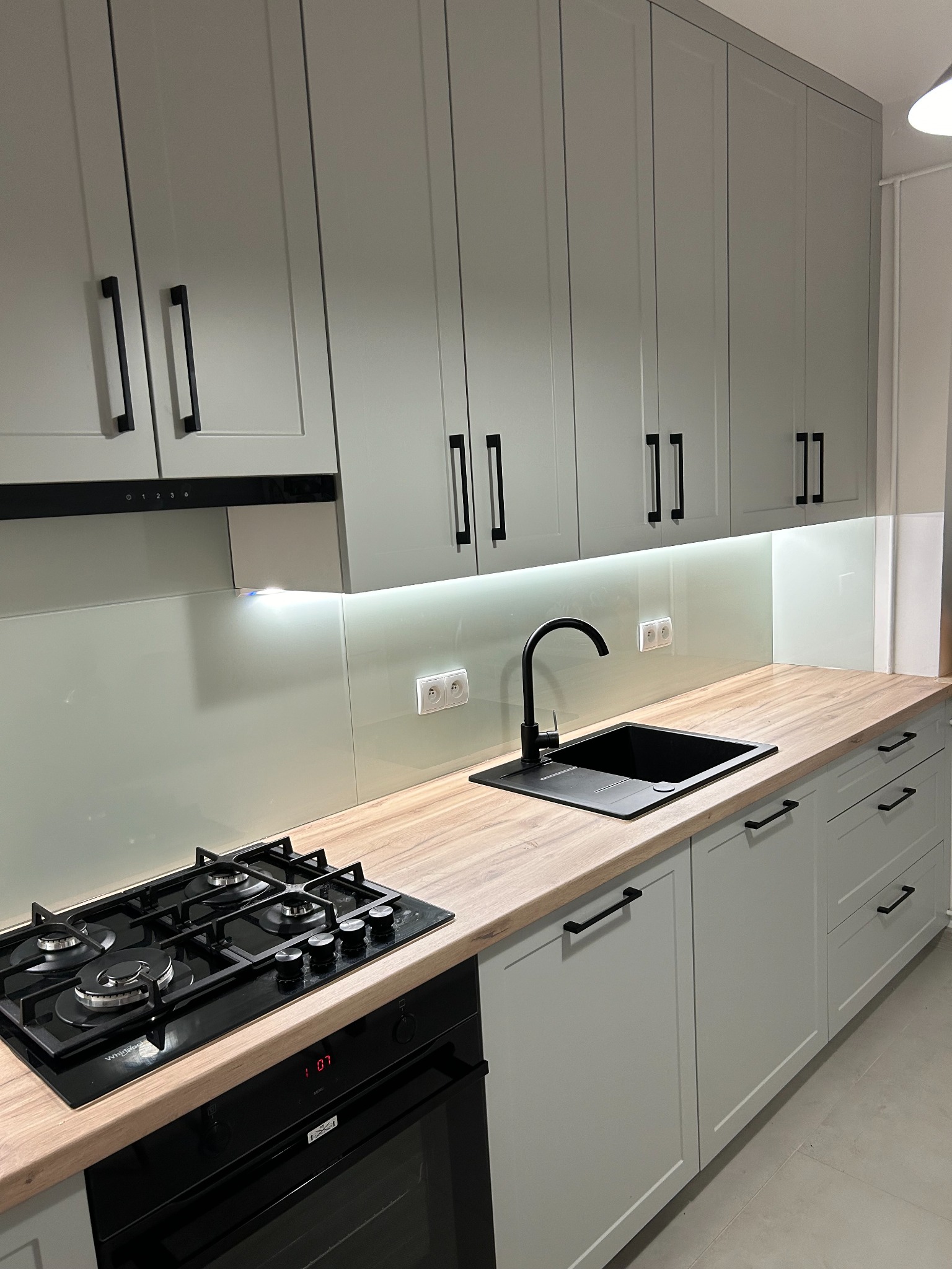 Szara kuchnia na wymiar z drewnianym blatem, czarną armaturą i płytą gazową, minimalistyczne uchwyty