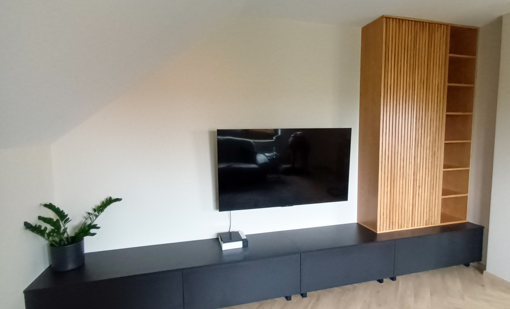 Zabudowa pod telewizor z szafkami w kolorze grafitowym i wysoką szafą z lamelami z jasnego drewna, uzupełniona rośliną doniczkową, pod skosem sufitu.