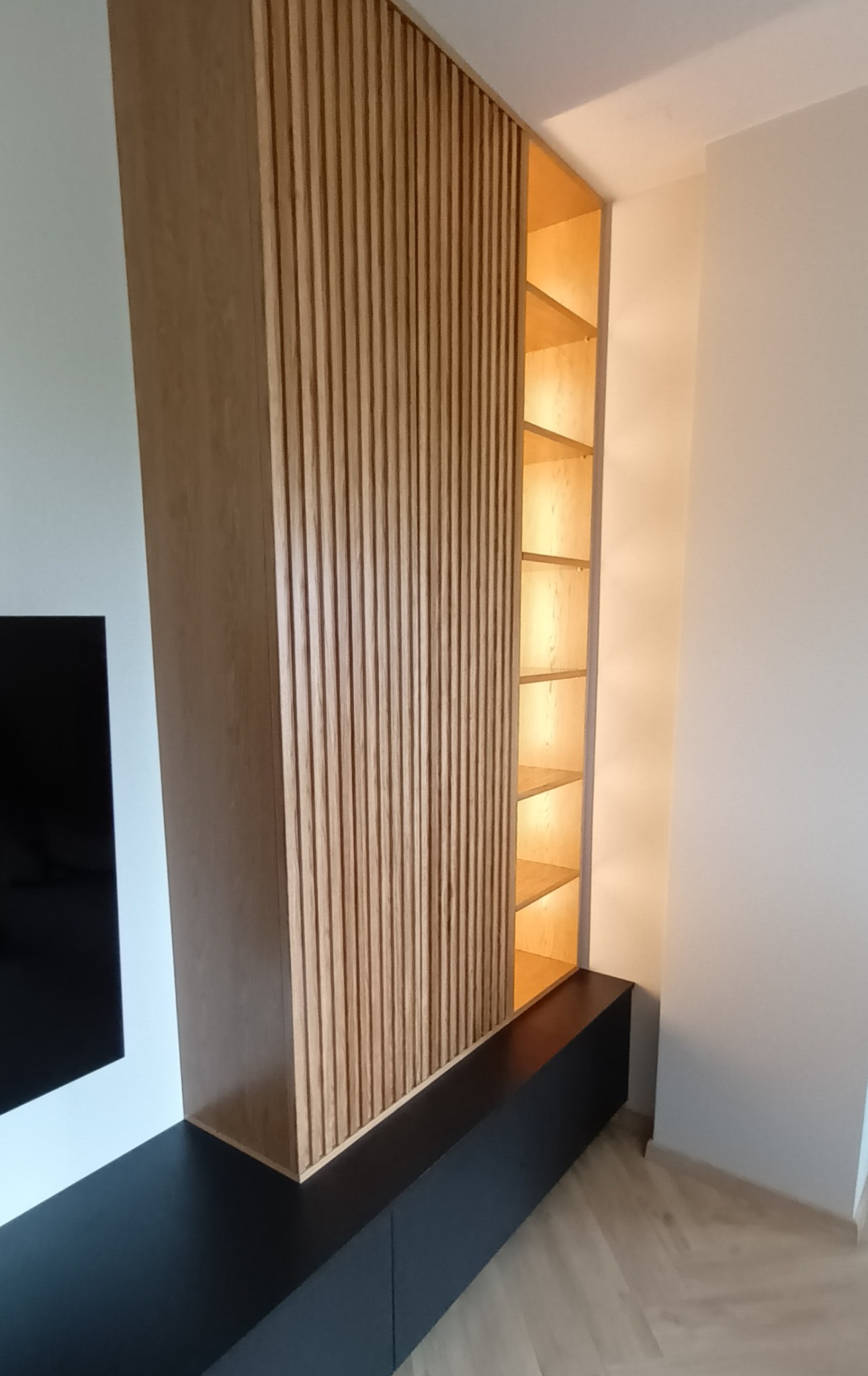 Nowoczesna szafa wnękowa z drewnianymi lamelami i podświetlanymi półkami, zintegrowana z czarnym panelem RTV.