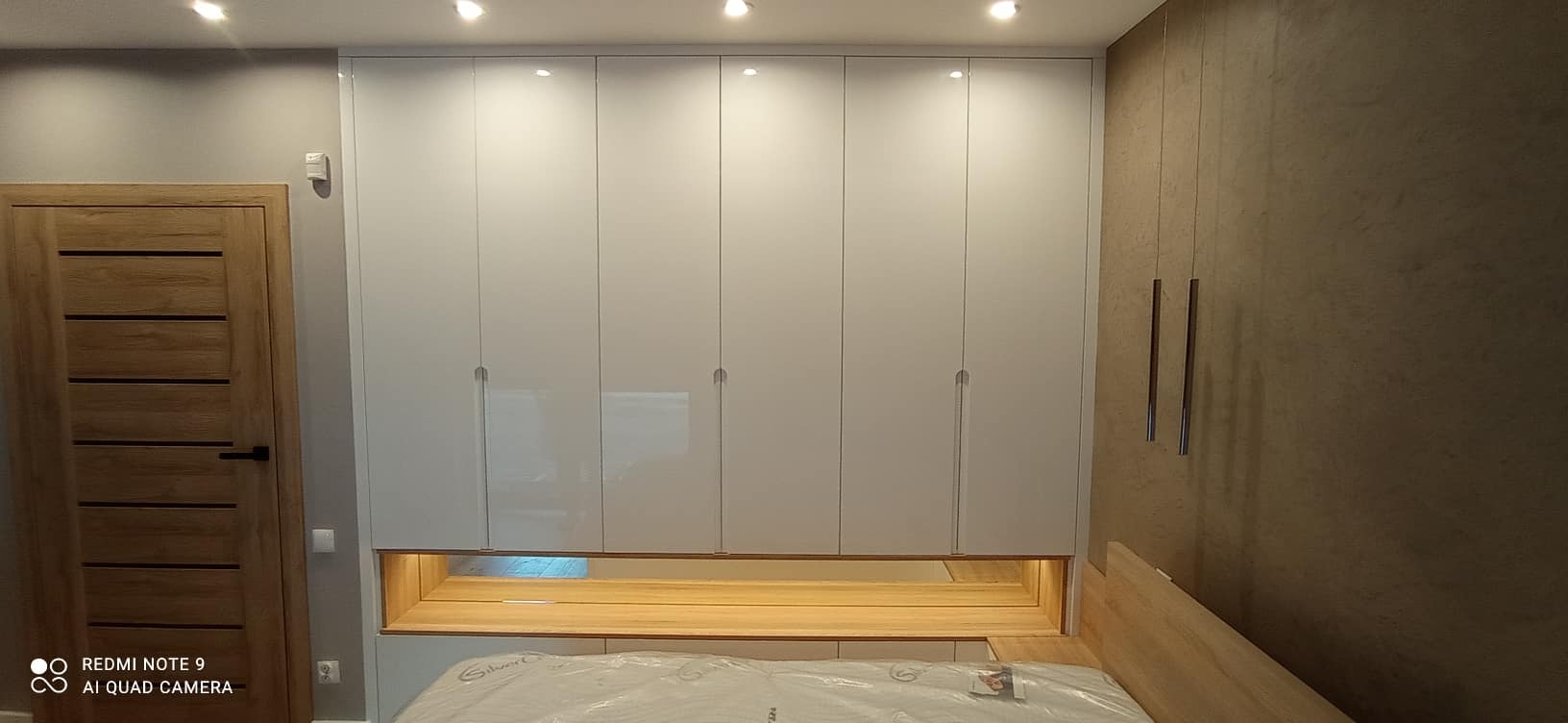 Biała szafa wnękowa z połyskiem, wkomponowana w aranżację sypialni nad łóżkiem, z drewnianą wnęką oświetloną od spodu, minimalistyczne uchwyty.