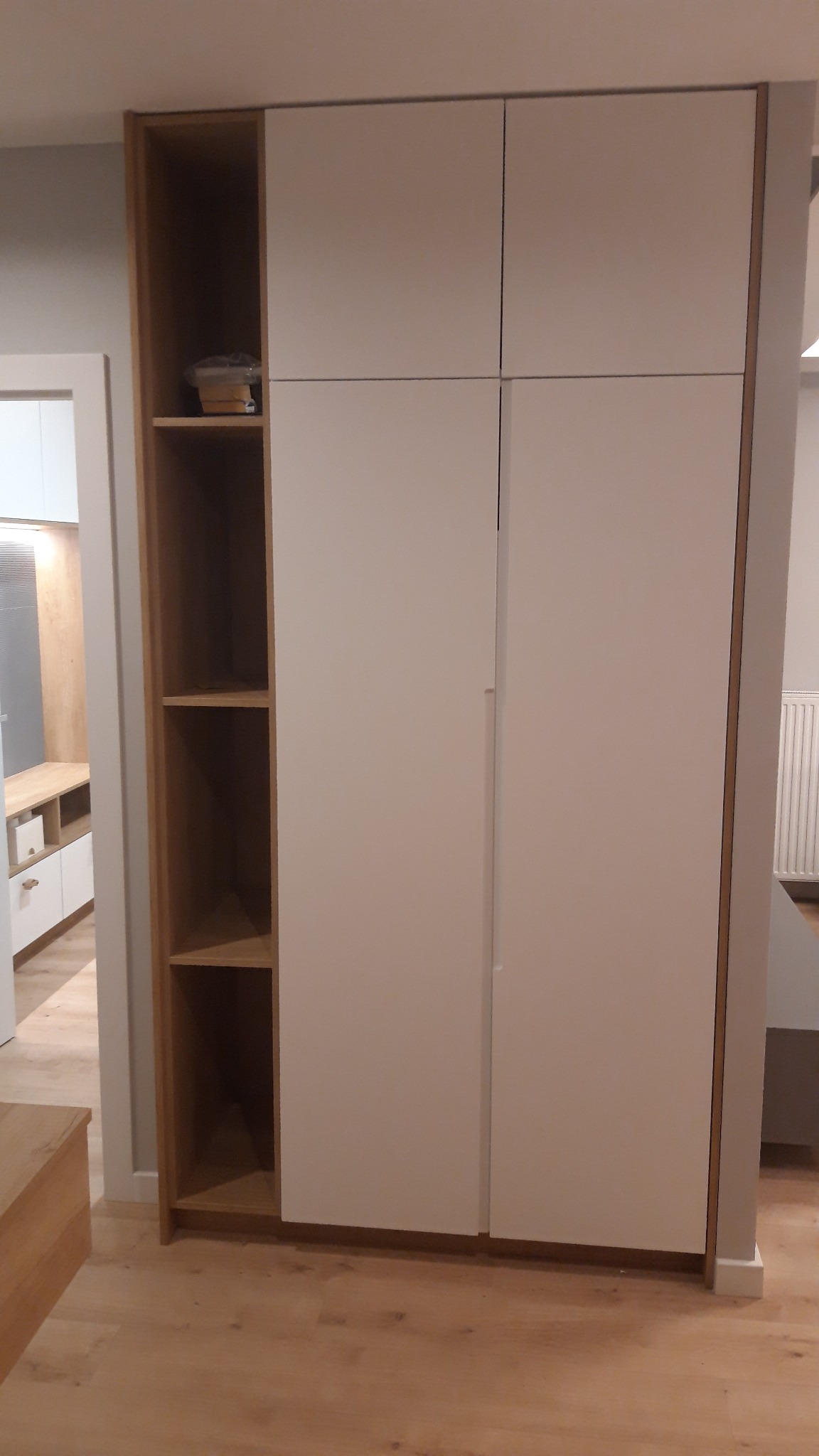 Biała szafa z przesuwnymi drzwiami i otwartymi półkami z jasnego drewna, widoczna na tle jasnego parkietu i fragmentu innego pomieszczenia.