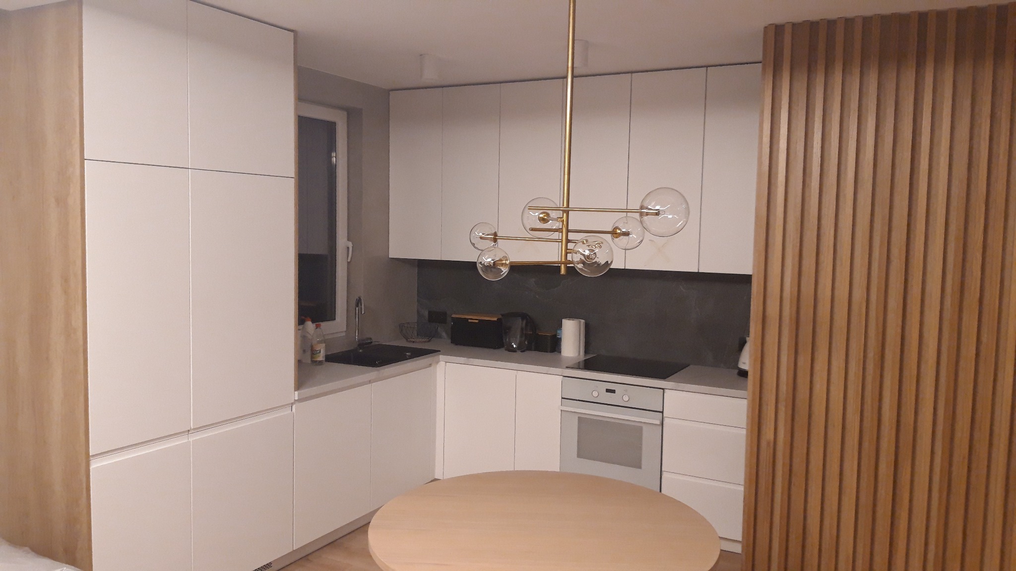 Nowoczesna kuchnia z białymi szafkami, okrągłym stołem i złotą lampą wiszącą, uzupełniona drewnianą ścianką działową z pionowych listew.