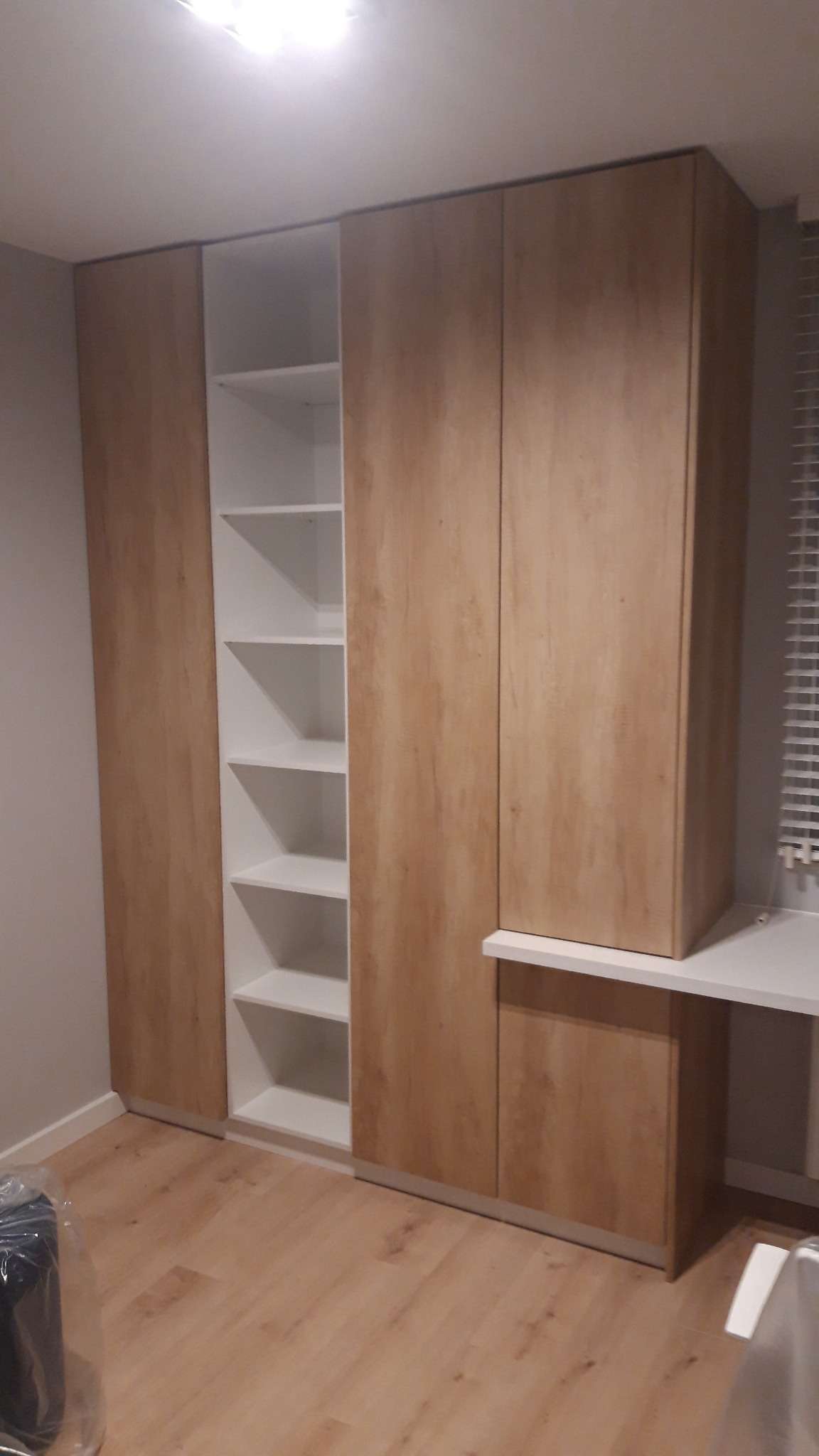 Wysoka szafa z drewnopodobnymi frontami i białymi półkami, wbudowana w ścianę, z blatem roboczym po prawej stronie, widok na wnętrze szafy z półkami.