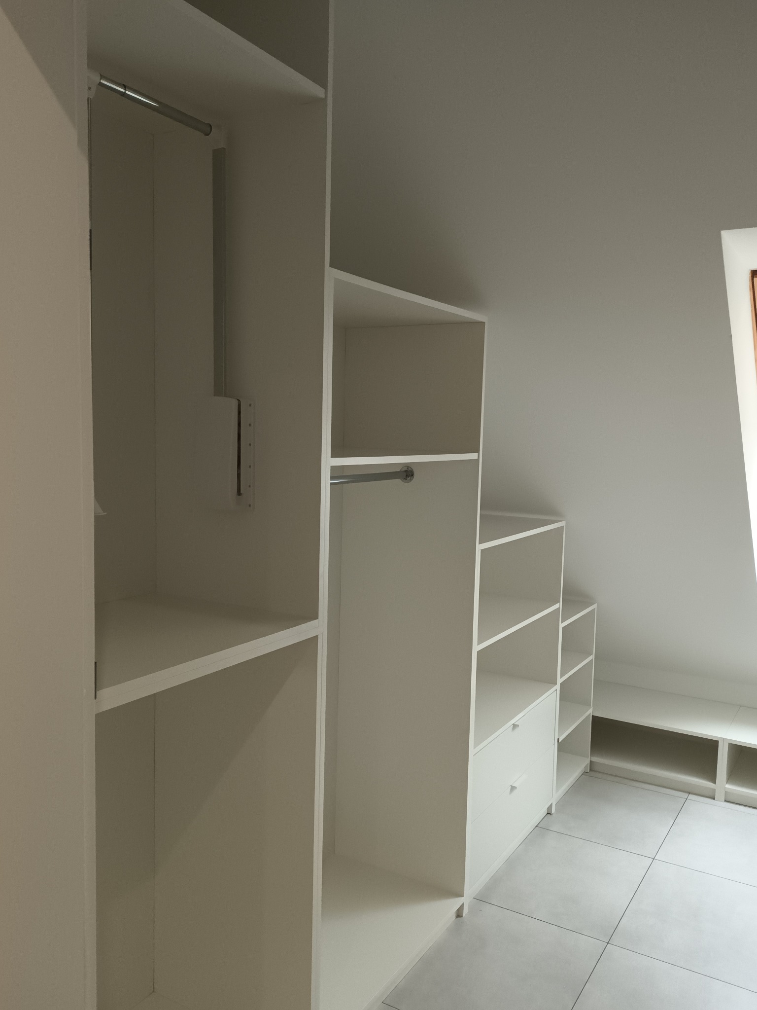 Biała zabudowa garderoby na poddaszu ze skośnym sufitem, widoczne półki, szuflady i drążki na ubrania, szara podłoga z płytek.