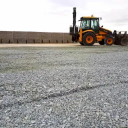 Żółta koparko-ładowarka Volvo na utwardzonej powierzchni z drobnego kruszywa, widoczny betonowy płot w tle.