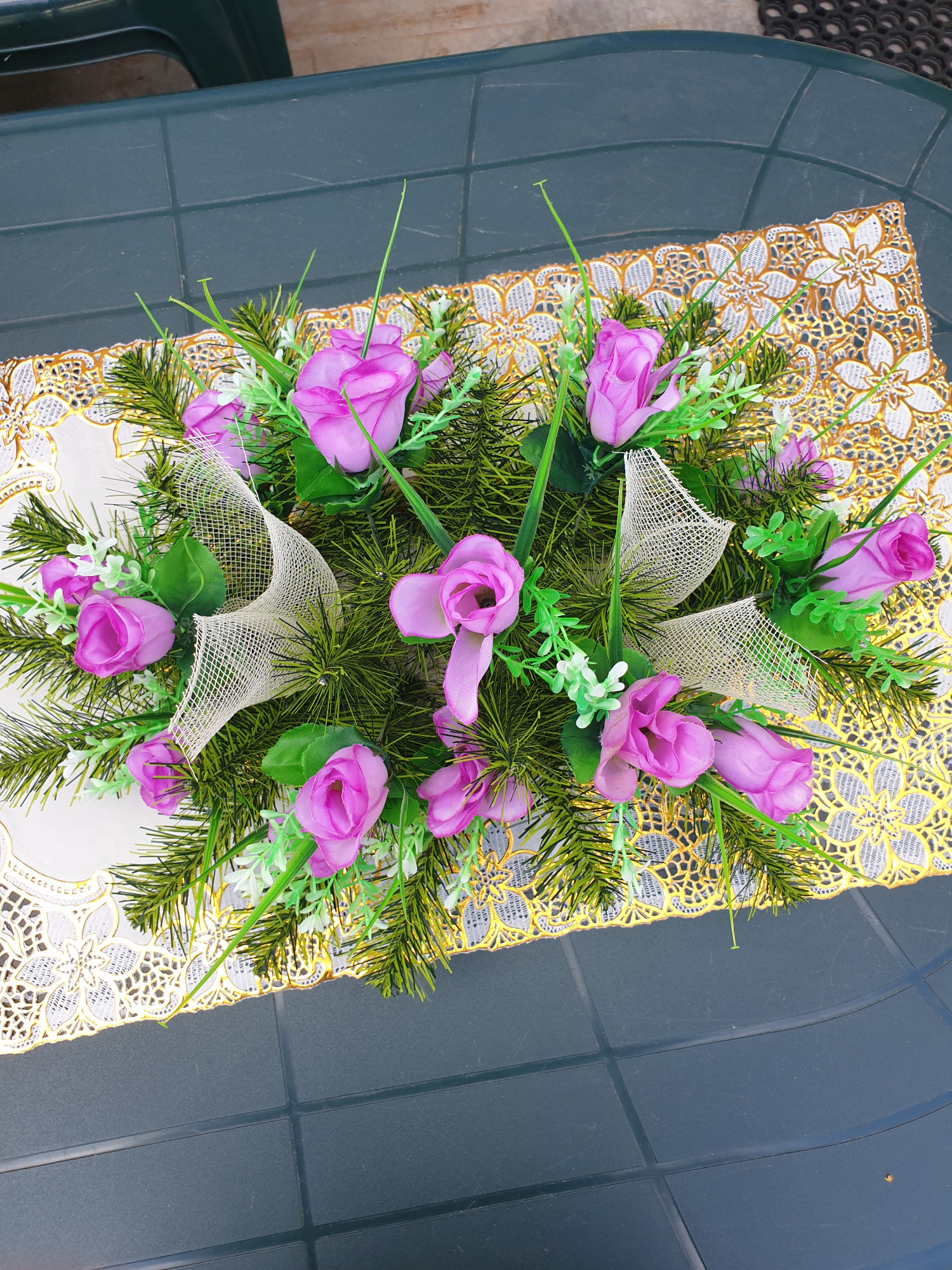 Dekoracyjny stroik z fioletowymi różami, zielonymi gałązkami i siatkową wstążką, umieszczony na białej serwecie ze złotym wzorem, leżącej na zielonym stole.