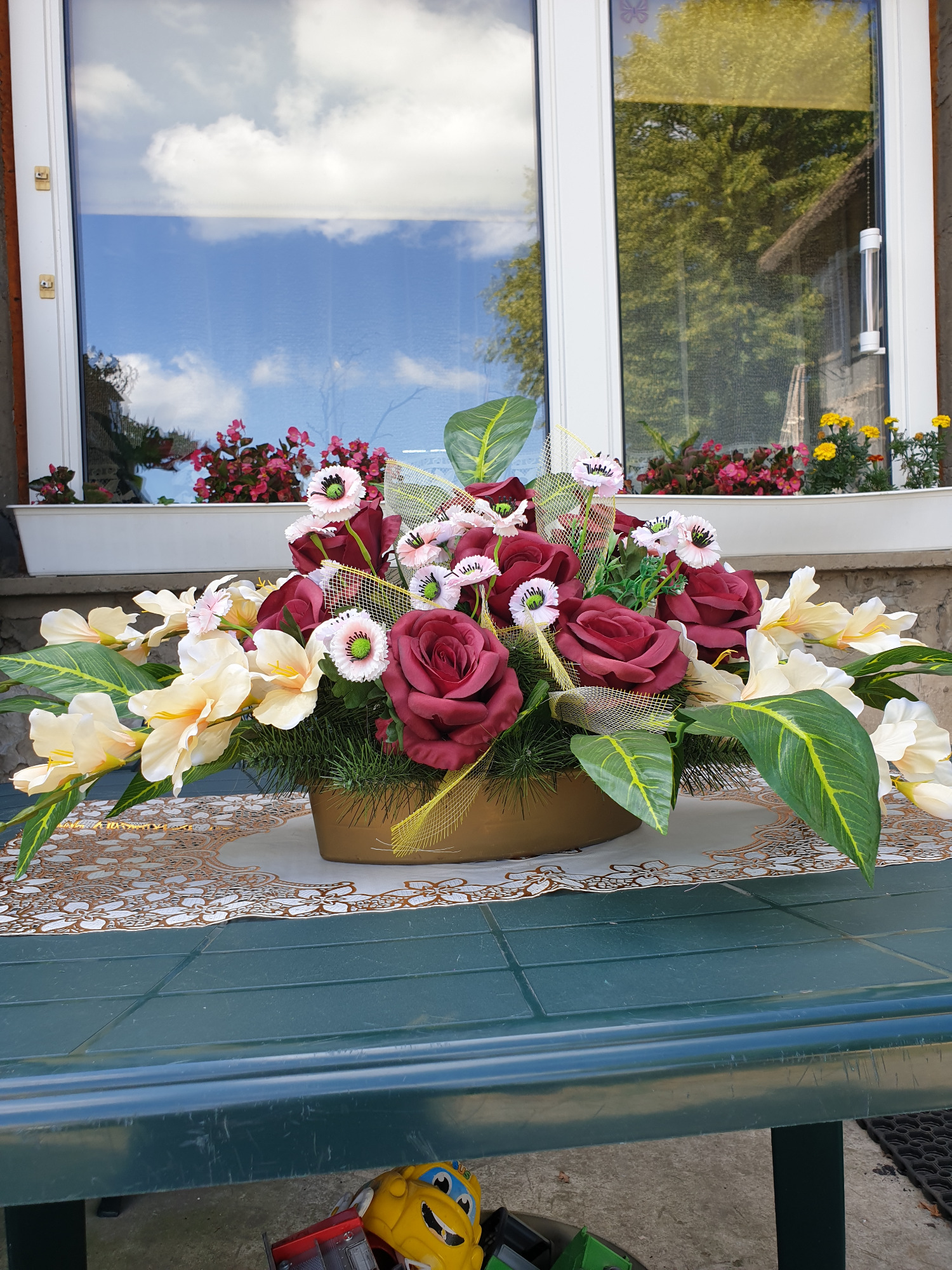 Sztuczny stroik z bordowych róż, białych irysów i maków w złotej siatce, ustawiony na stole przed oknem z widokiem na niebo i kwiaty w doniczce.