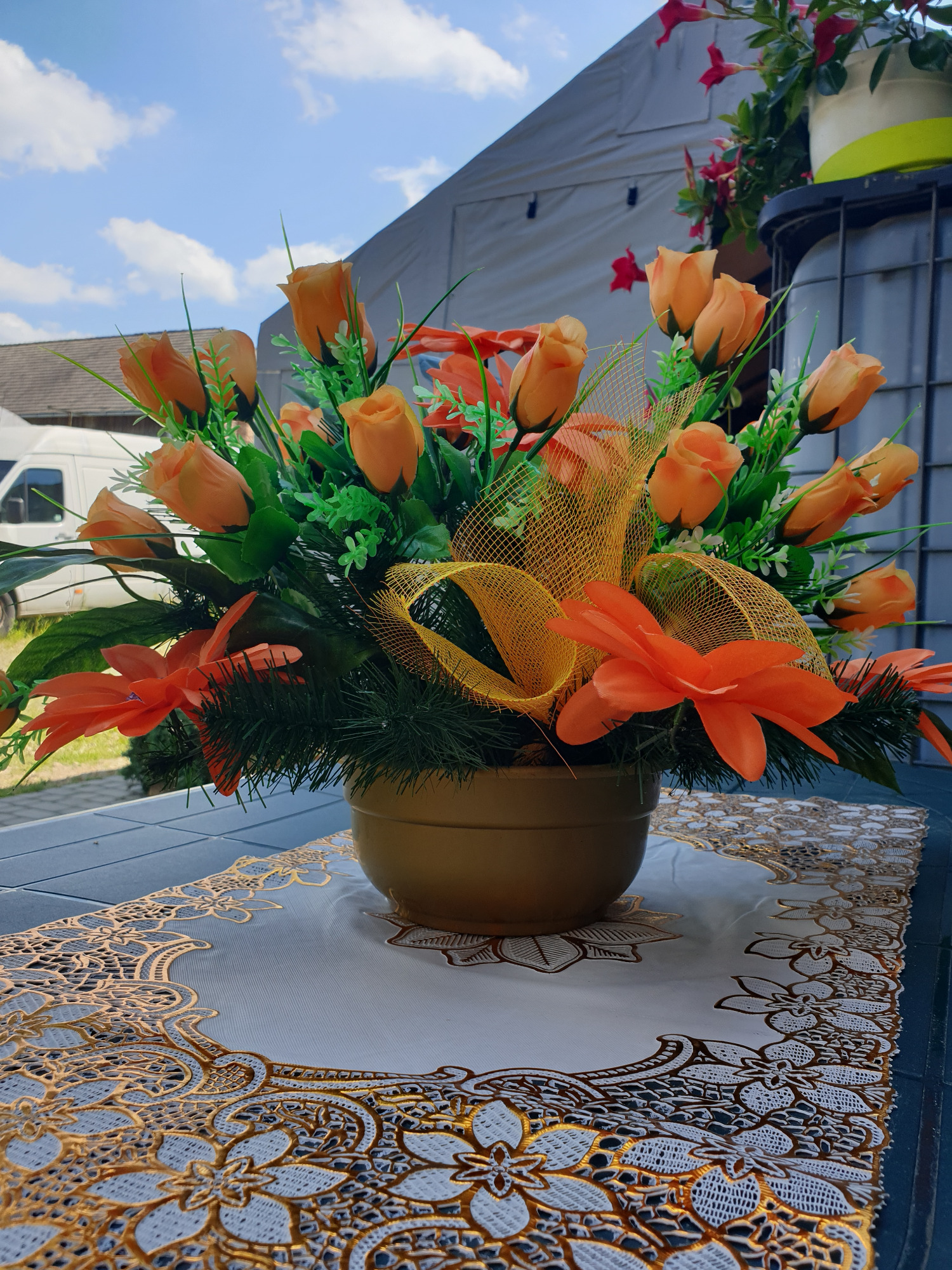 Sztuczny stroik z pomarańczowymi różami i wstążkami w złotym wazonie, ustawiony na białym obrusie z ażurowym, złotym wzorem.