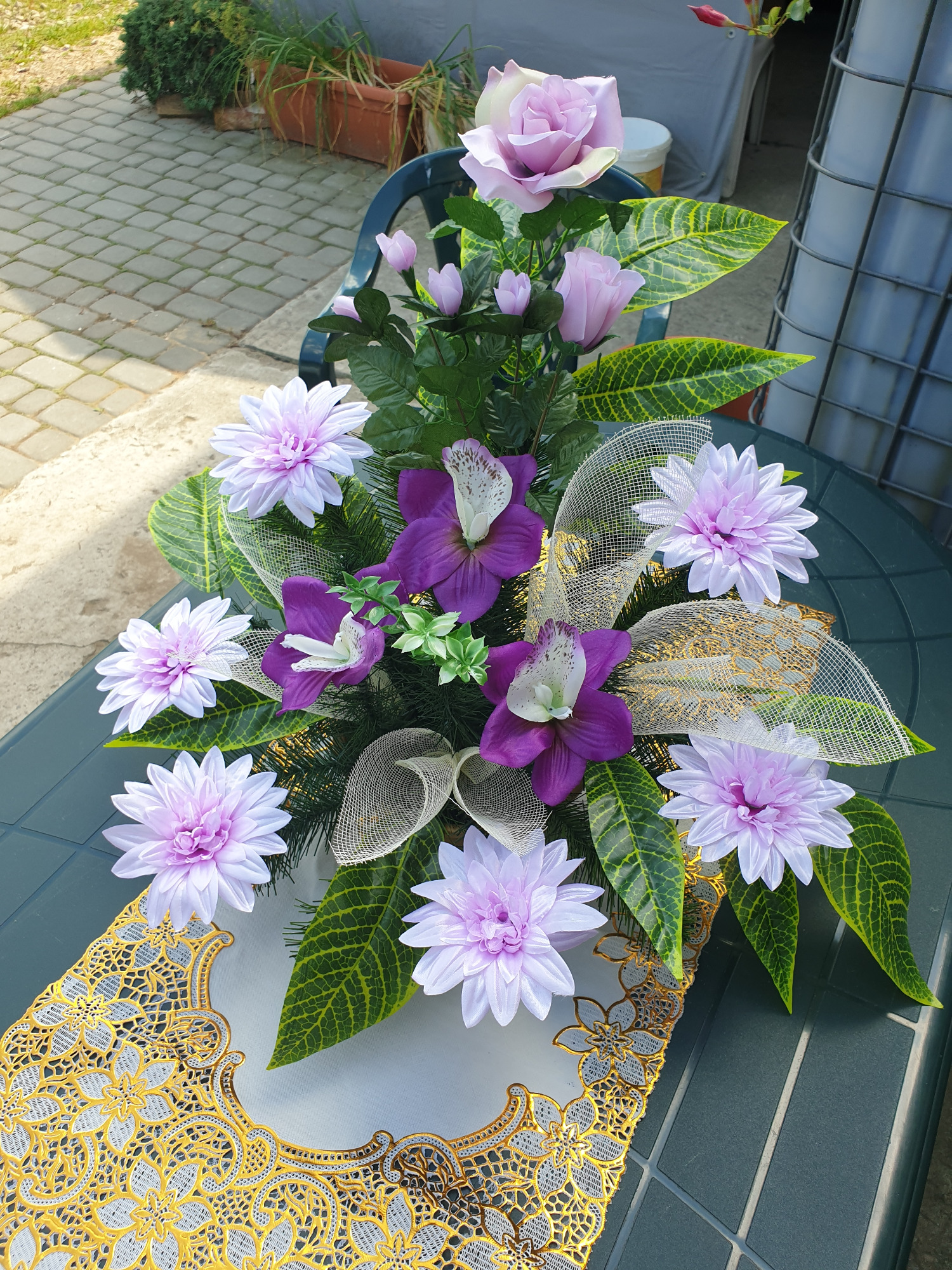 Kompozycja kwiatowa ze sztucznych kwiatów w odcieniach fioletu i bieli, z dekoracyjną siatką i złotą serwetką, umieszczona na zielonym stole.