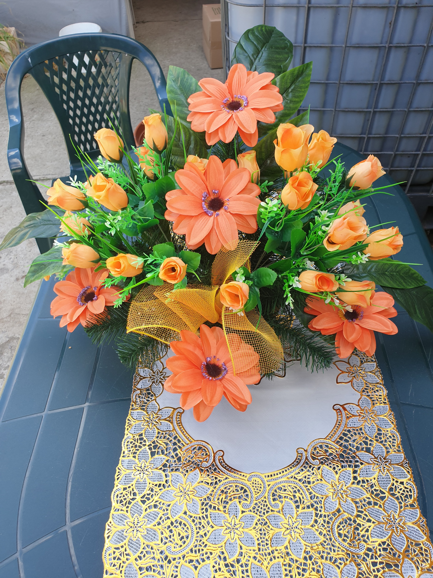 Kompozycja ze sztucznych pomarańczowych kwiatów i różyczek, ozdobiona złotą siatką, na stole z dekoracyjną serwetą w Poznaniu. W tle widoczne krzesło.