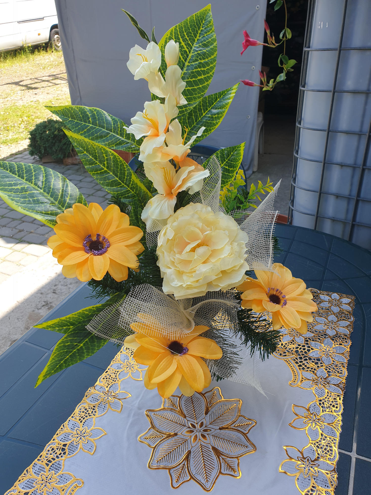Kompozycja ze sztucznych kwiatów w odcieniach bieli i żółci, z gerberami i irysami, ozdobiona tiulową siatką, umieszczona na stole przykrytym białym obrusem z ażurowym, złotym wzorem.