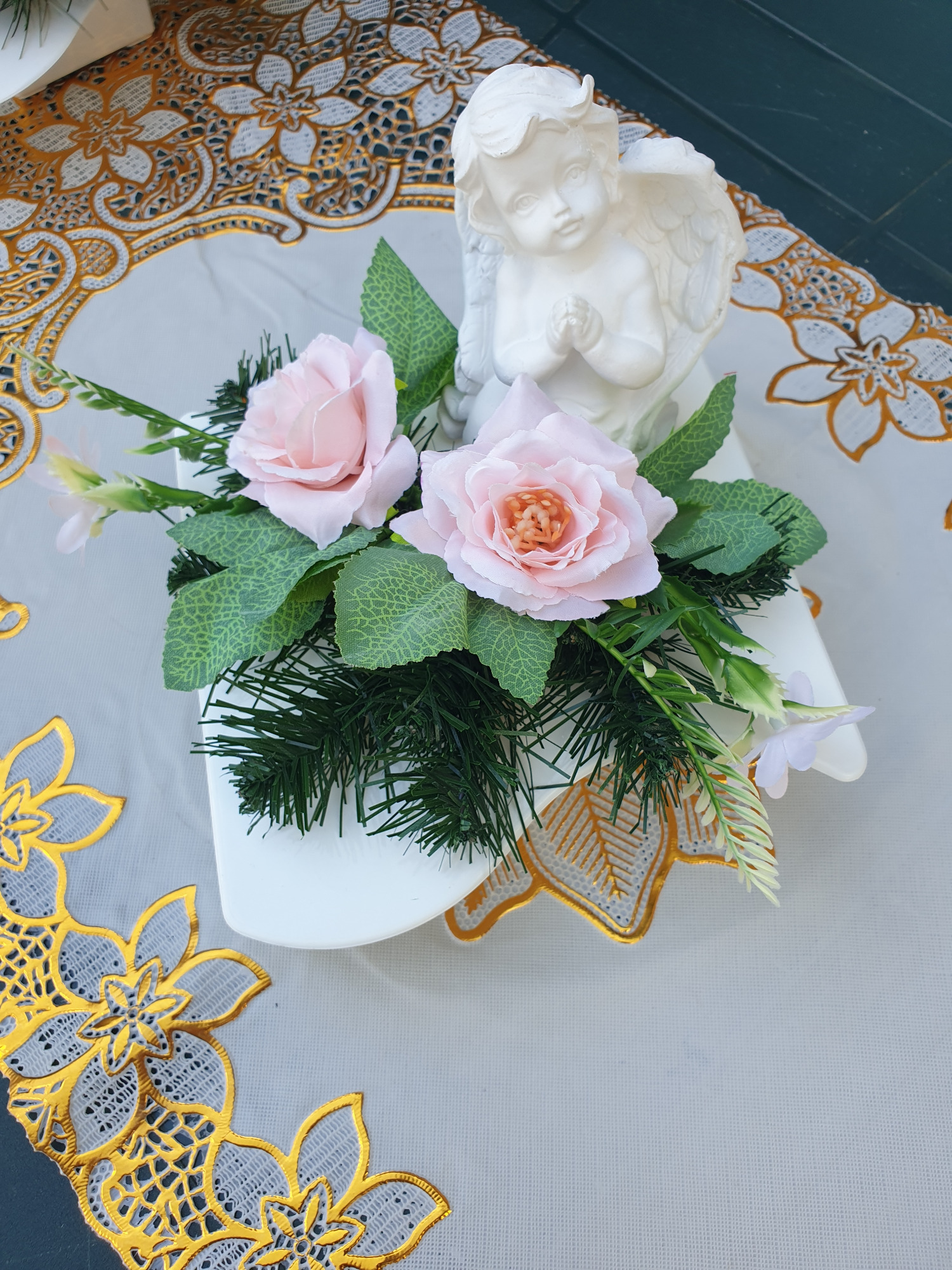 Dekoracja stołu z białym aniołkiem, dwoma różowymi różami, zielonymi liśćmi i igliwiem na białym podkładzie z haftowanymi złotymi kwiatami.
