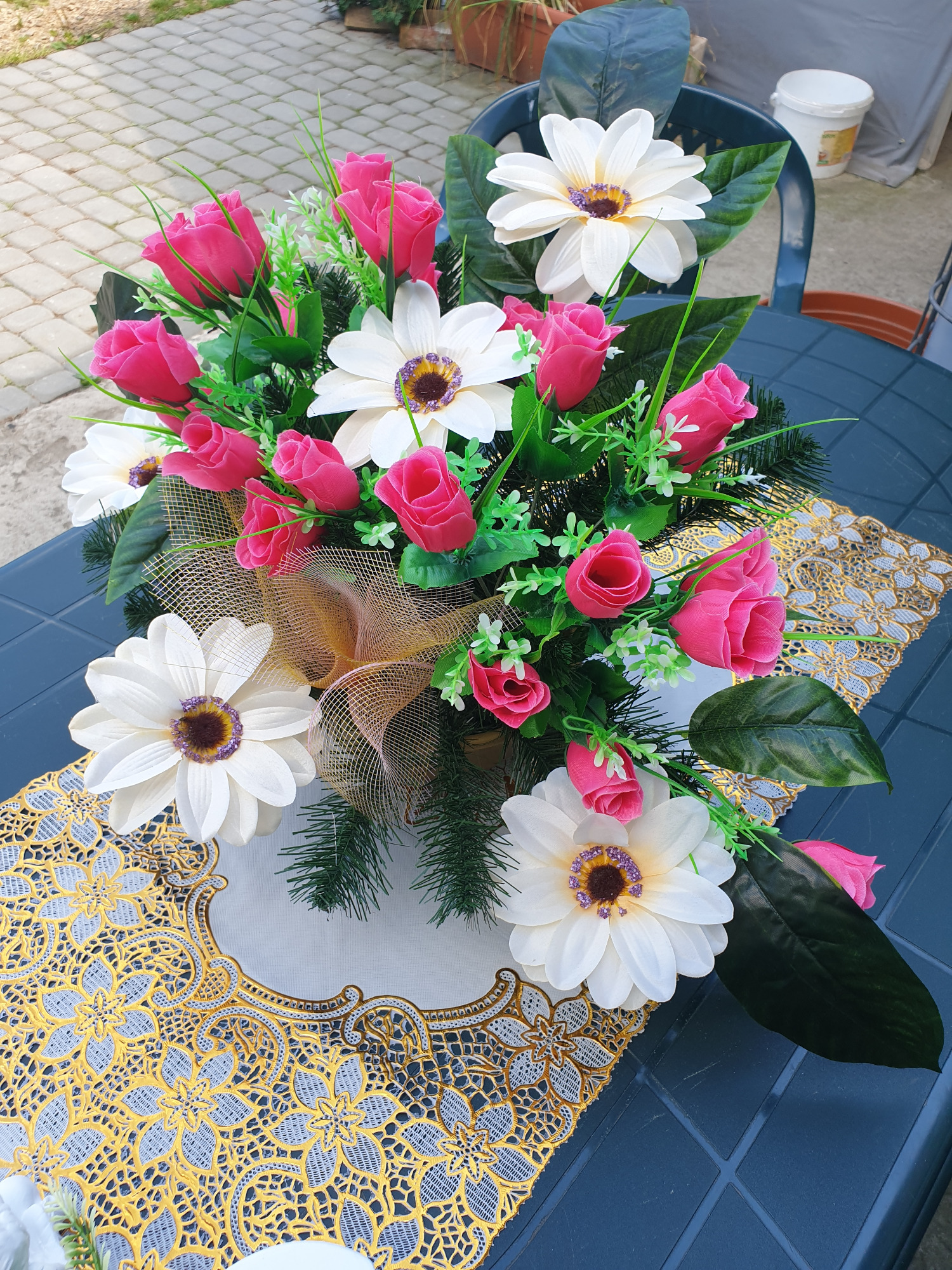 Kompozycja kwiatowa ze sztucznych różowych róż i białych gerber na ozdobnej, złotej serwecie na niebieskim stole.