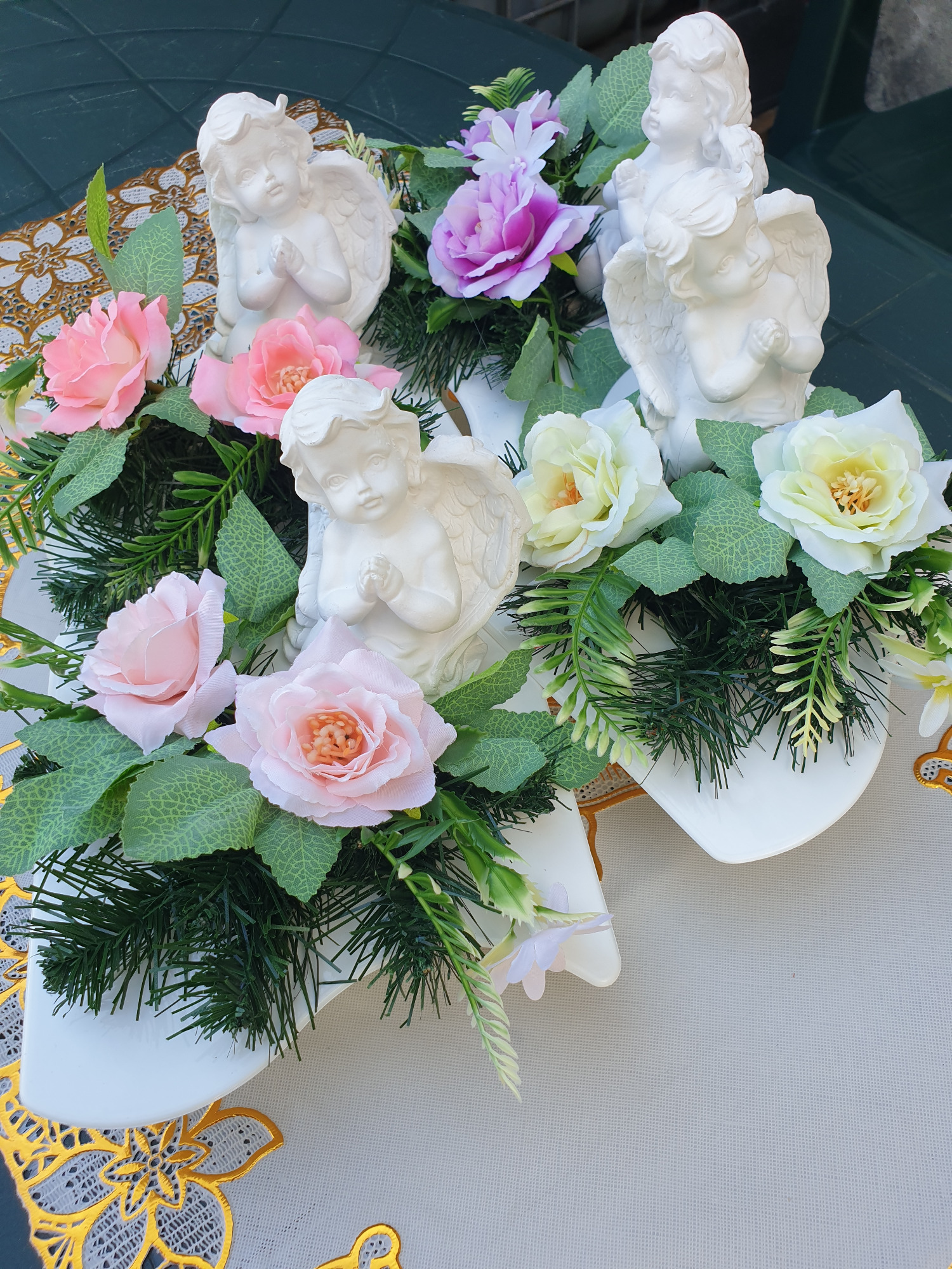 Kompozycja kwiatowa z białymi figurkami aniołów na białych podstawkach, z różowymi, białymi i fioletowymi różami oraz zielonymi gałązkami, na stole z ażurową, złotą serwetą.