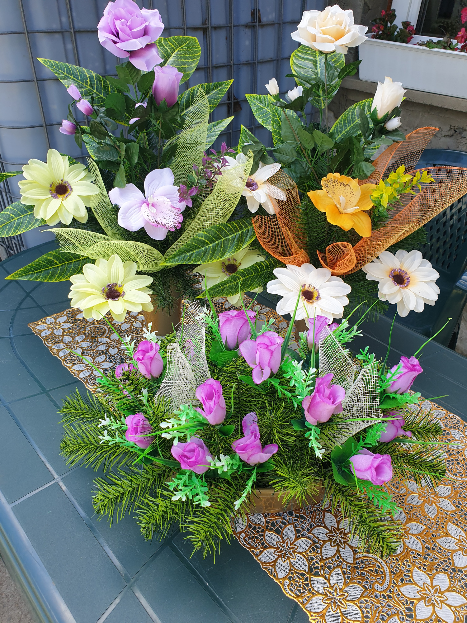 Kompozycje kwiatowe ze sztucznych kwiatów w odcieniach fioletu, żółci i bieli, ozdobione siatką, ustawione na złotej serwecie na zielonym stole.