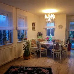 Jadalnia w okresie świątecznym: białe, zwiewne zasłony z dekoracyjnymi koralikami w oknach, drewniany stół nakryty świątecznym obrusem, mała choinka, drewniana podłoga i dywan w kwiaty.