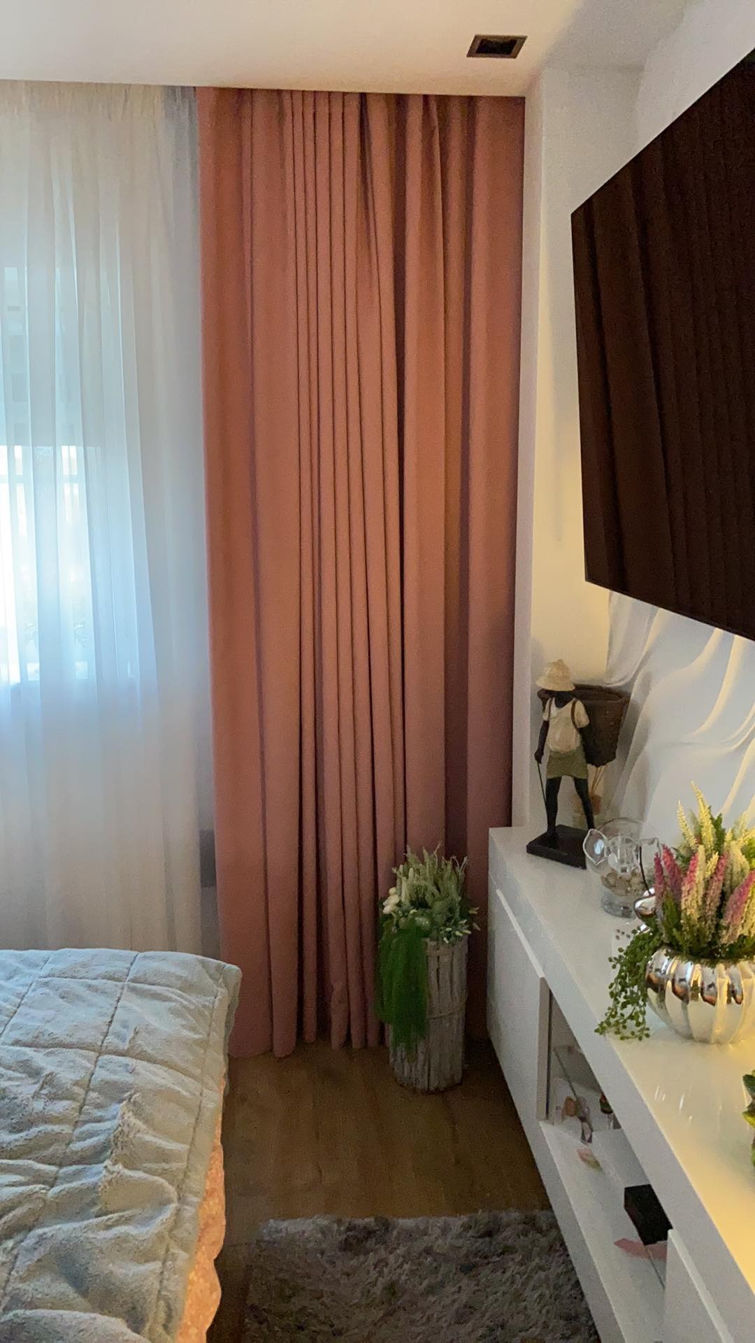 Fragment sypialni z zasłoniętym oknem: biała firanka i pudrowo-różowa, gruba zasłona, dekoracje na białej komodzie pod telewizorem.