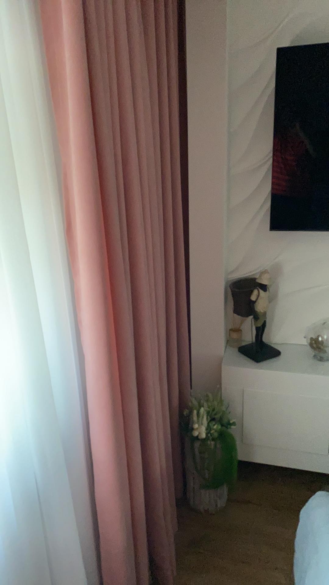 Delikatne, plisowane zasłony w odcieniach bieli i pudrowego różu, częściowo zasłaniające okno, z dekoracyjnym bukietem kwiatów w wazonie i elementami dekoracyjnymi na szafce w tle.