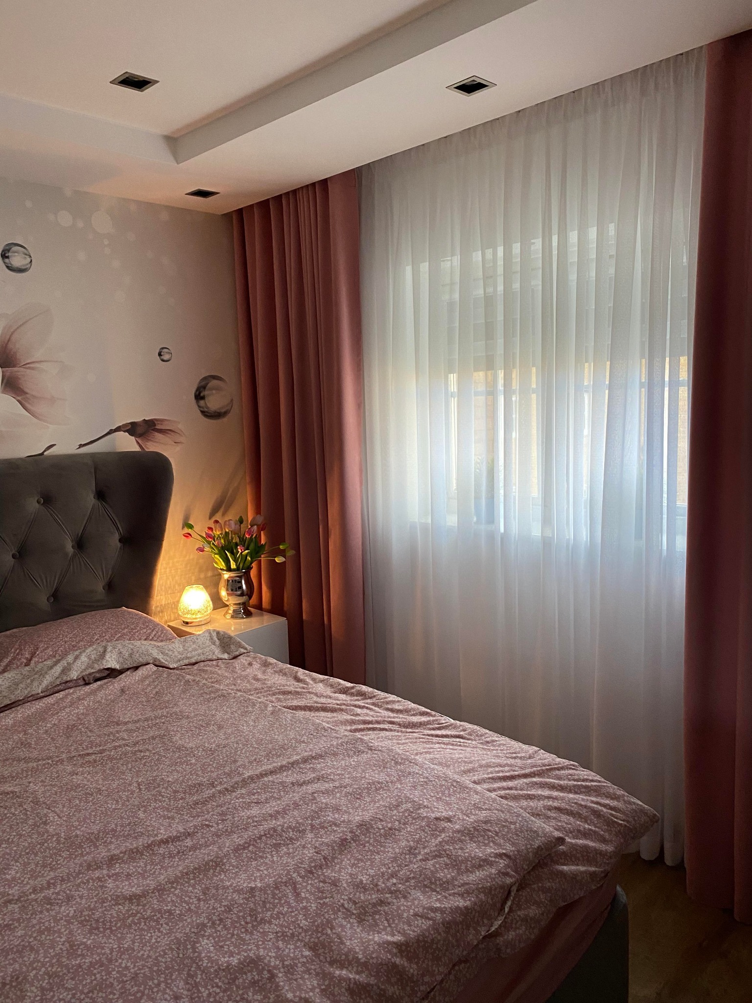 Sypialnia z zasłonami w odcieniach różu: delikatna firanka przepuszcza światło, a grube zasłony po bokach dodają przytulności. Na szafce nocnej stoi wazon z tulipanami obok świecącej lampki.