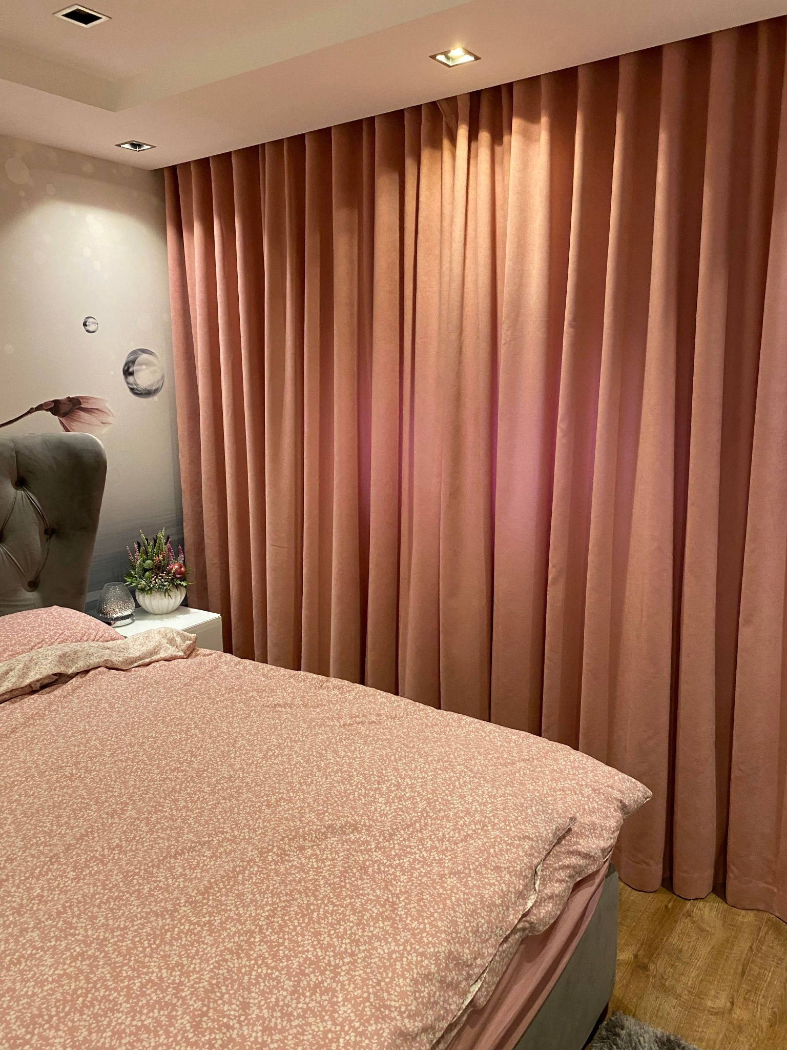 Różowe, plisowane zasłony w sypialni z łóżkiem w kwiatowy wzór i tapetą z motywem kwiatowym, oświetlone punktowo.