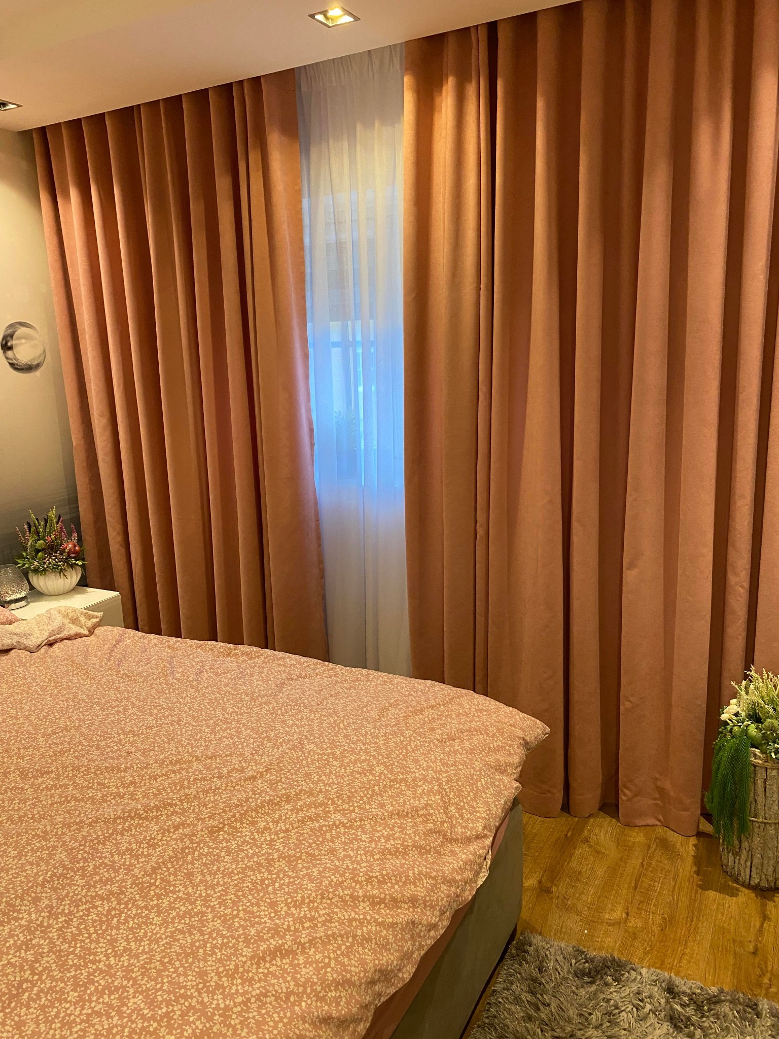 Beżowe, grube zasłony zaciemniające w sypialni, z widocznym fragmentem łóżka z narzutą w drobne kwiatki i dekoracjami na szafce nocnej oraz podłodze.