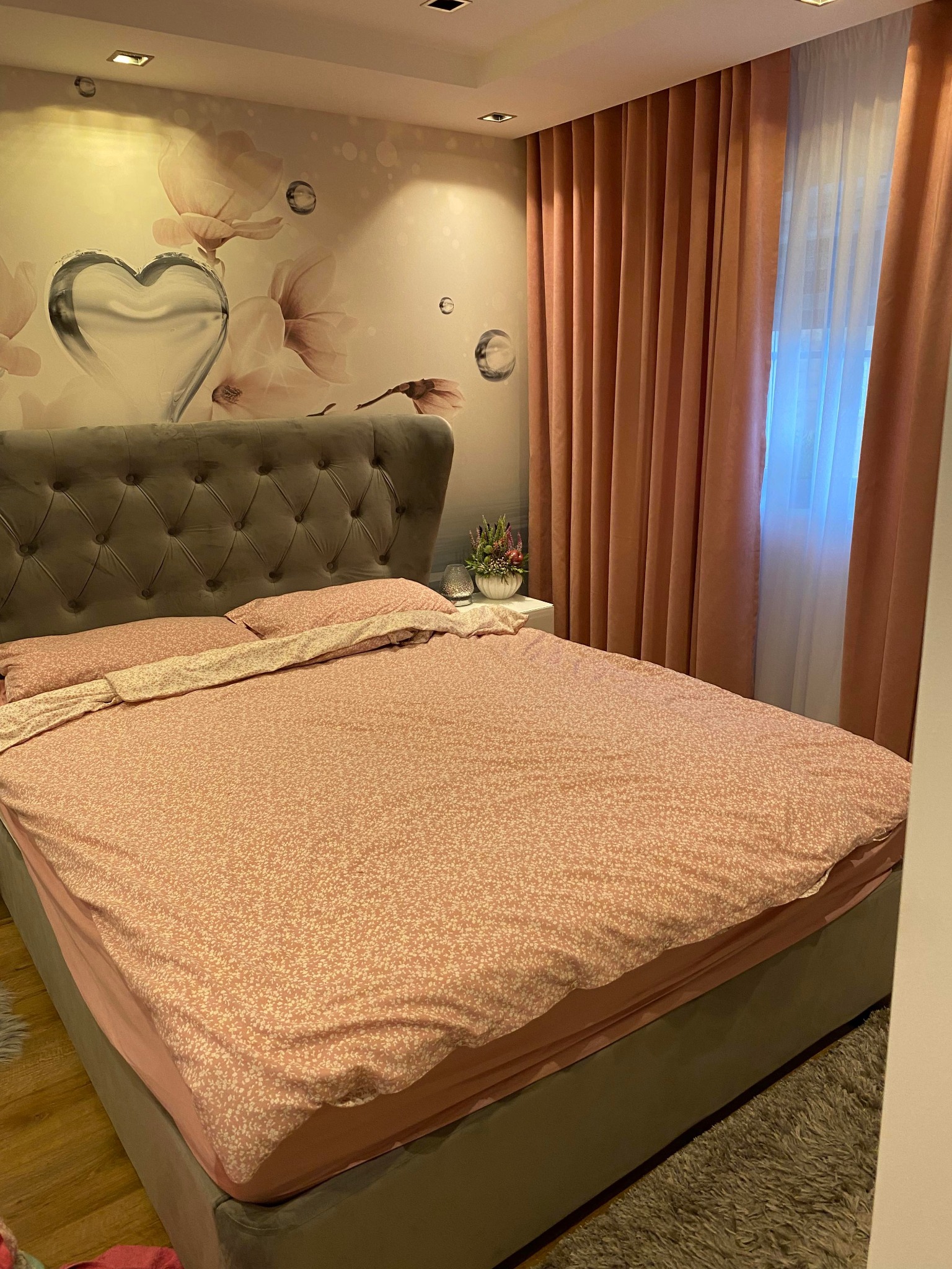 Sypialnia z łóżkiem z szarym, pikowanym zagłówkiem, przykrytym różową pościelą w drobne kwiatki; na ścianie tapeta z motywem kwiatowym i sercem; okno zasłonięte długimi, różowymi zasłonami.