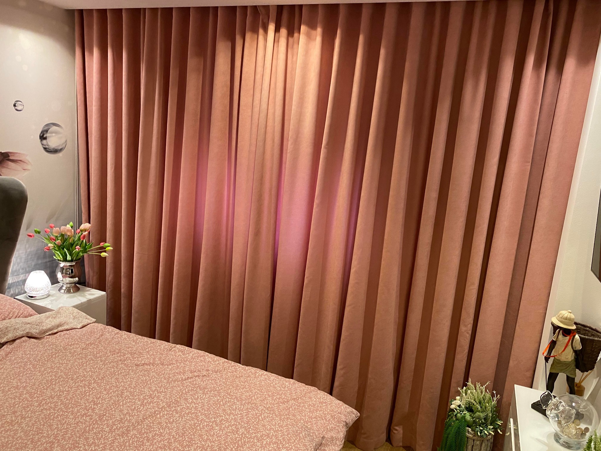 Pokój z zasłoniętym oknem, zasłony w odcieniu pudrowego różu, dopasowana pościel na łóżku, bukiet tulipanów w srebrnym wazonie na szafce nocnej, dekoracyjna figurka.