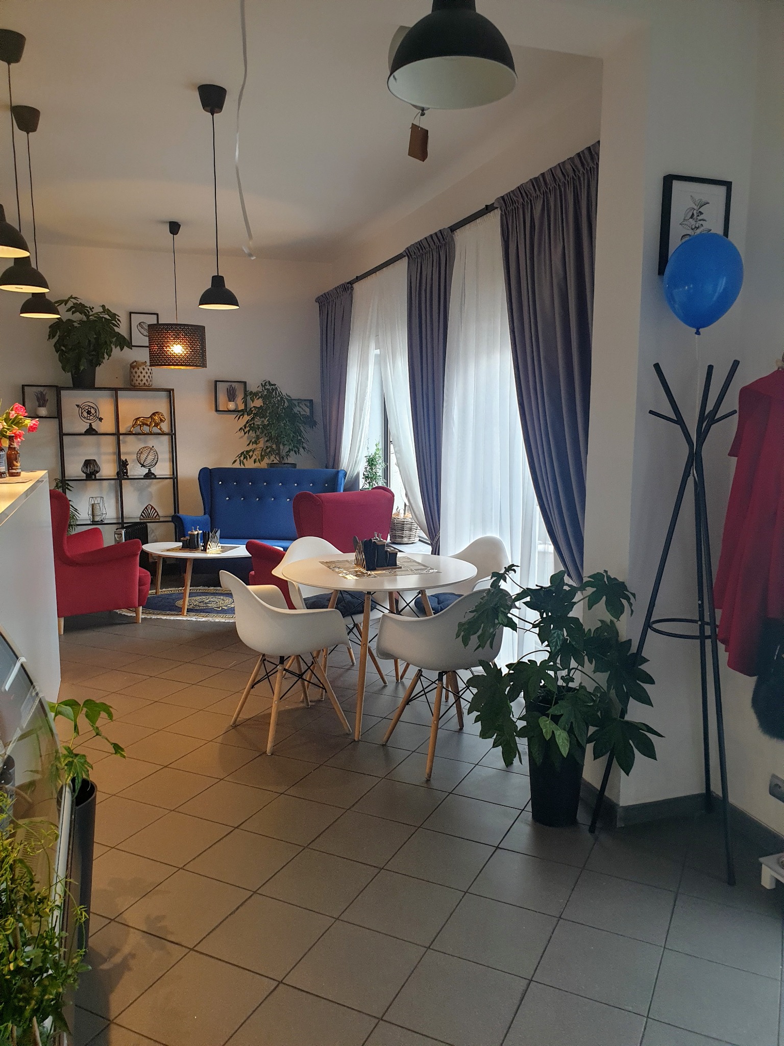 Wnętrze kawiarni lub restauracji z aranżacją okna: szare zasłony z białymi firanami, okrągłe stoły z białymi krzesłami, niebieska sofa i czerwone fotele, oświetlenie punktowe i wiszące lampy.