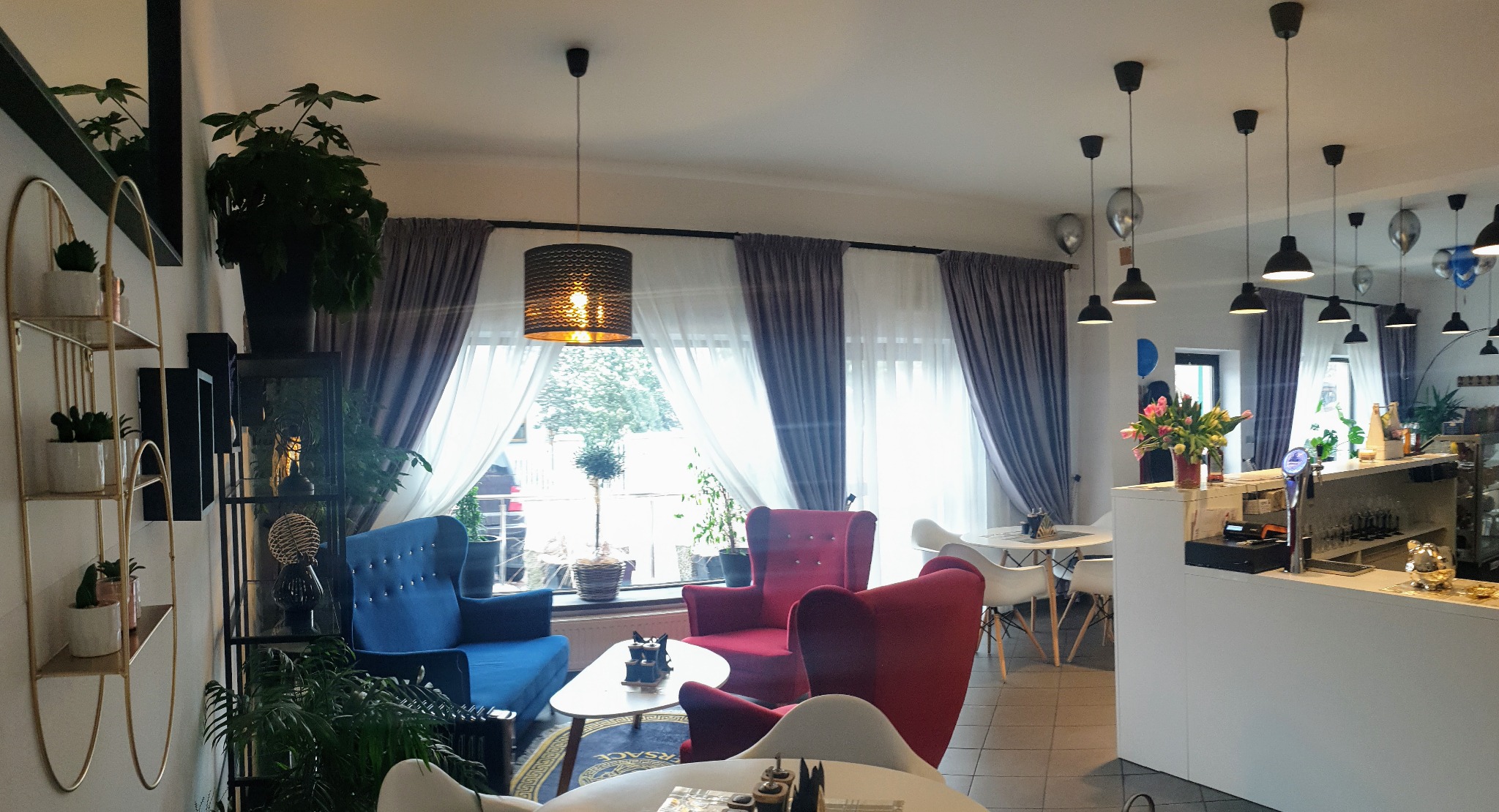 Wnętrze kawiarni z zasłonami w odcieniach szarości i bieli, niebieskim i czerwonym fotelem, widok z perspektywy klienta.