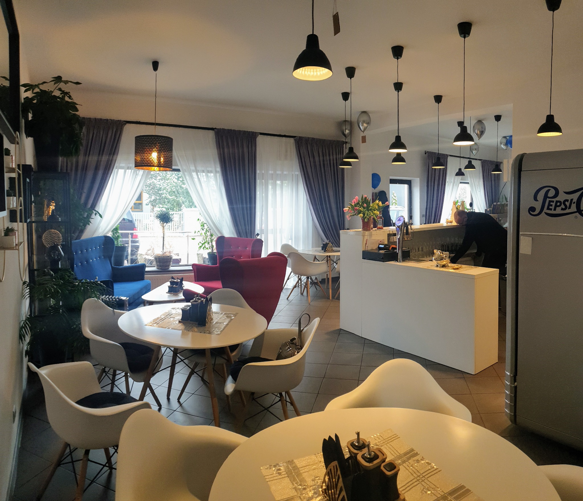 Wnętrze kawiarni z zasłonami w oknach, wiszącymi lampami i różnokolorowymi fotelami.