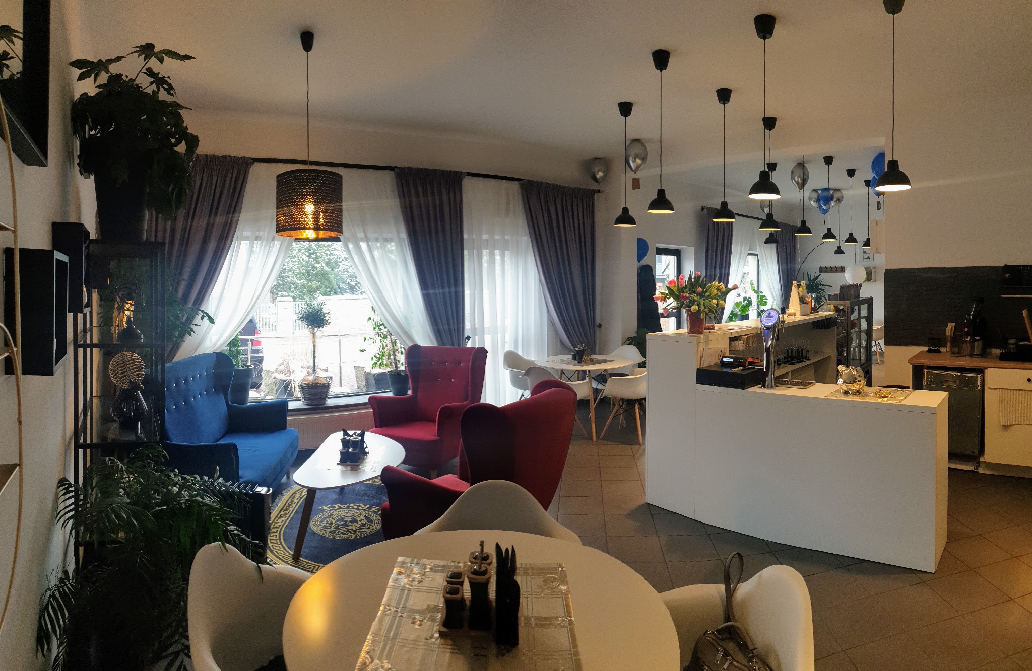 Przestronne wnętrze kawiarni z zasłonami w oknach, lampami wiszącymi, roślinami doniczkowymi i meblami w stylu retro. Widoczne niebieskie i czerwone fotele obite aksamitem, białe stoliki i krzesła...