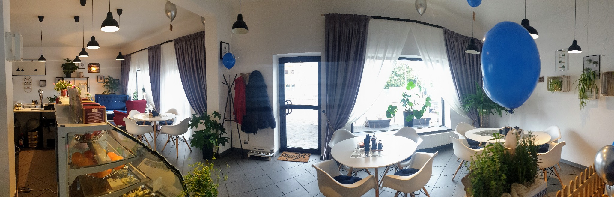 Wnętrze kawiarni z dużymi oknami, przez które widać zieleń, zasłoniętych częściowo szarymi i białymi zasłonami. Białe stoliki z krzesłami w stylu skandynawskim, dekoracje roślinne i balony tworzą...