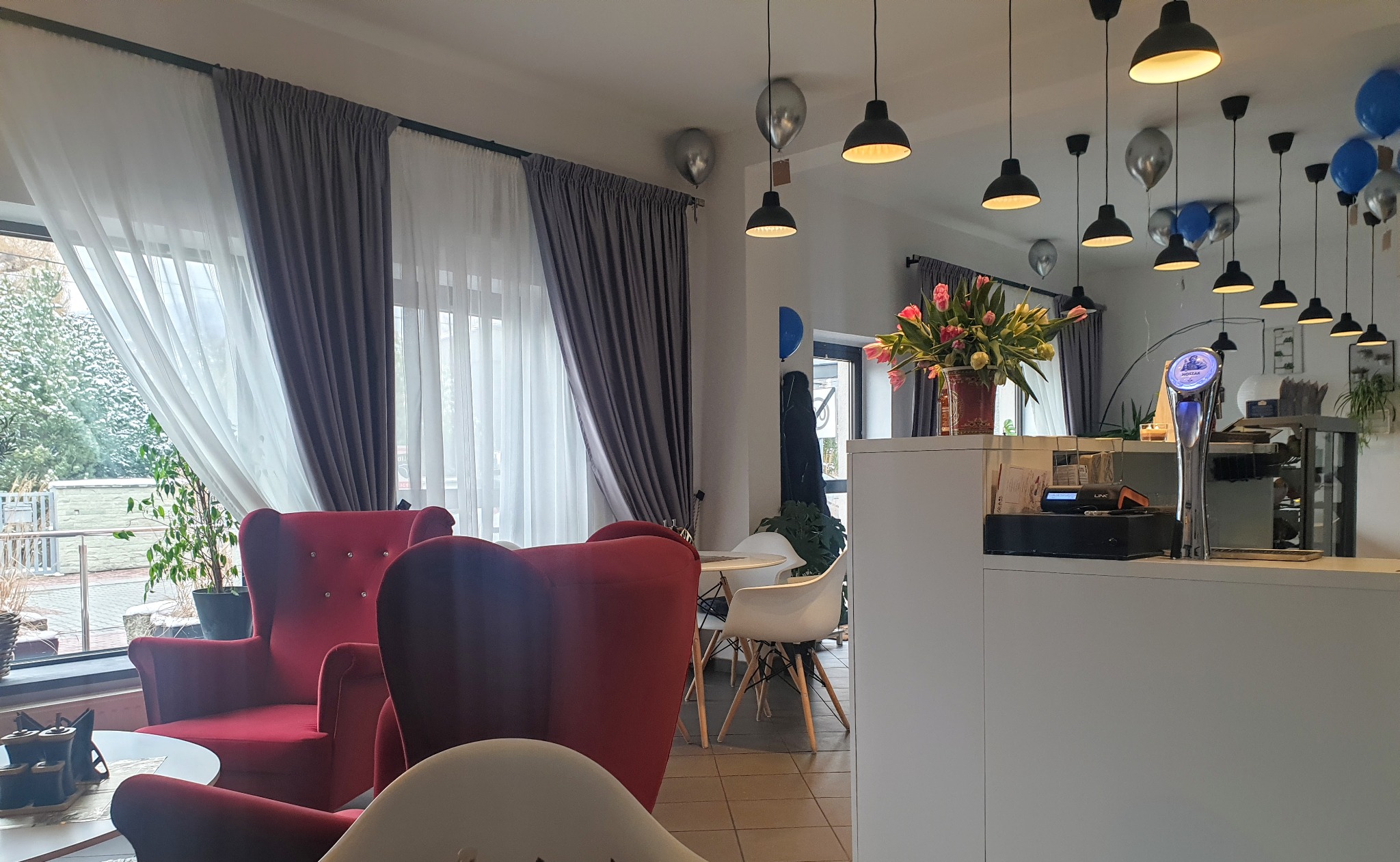 Wnętrze kawiarni z czerwonymi fotelami klubowymi przy oknie z szarymi i białymi zasłonami, dekorowane balonami i wiszącymi lampami