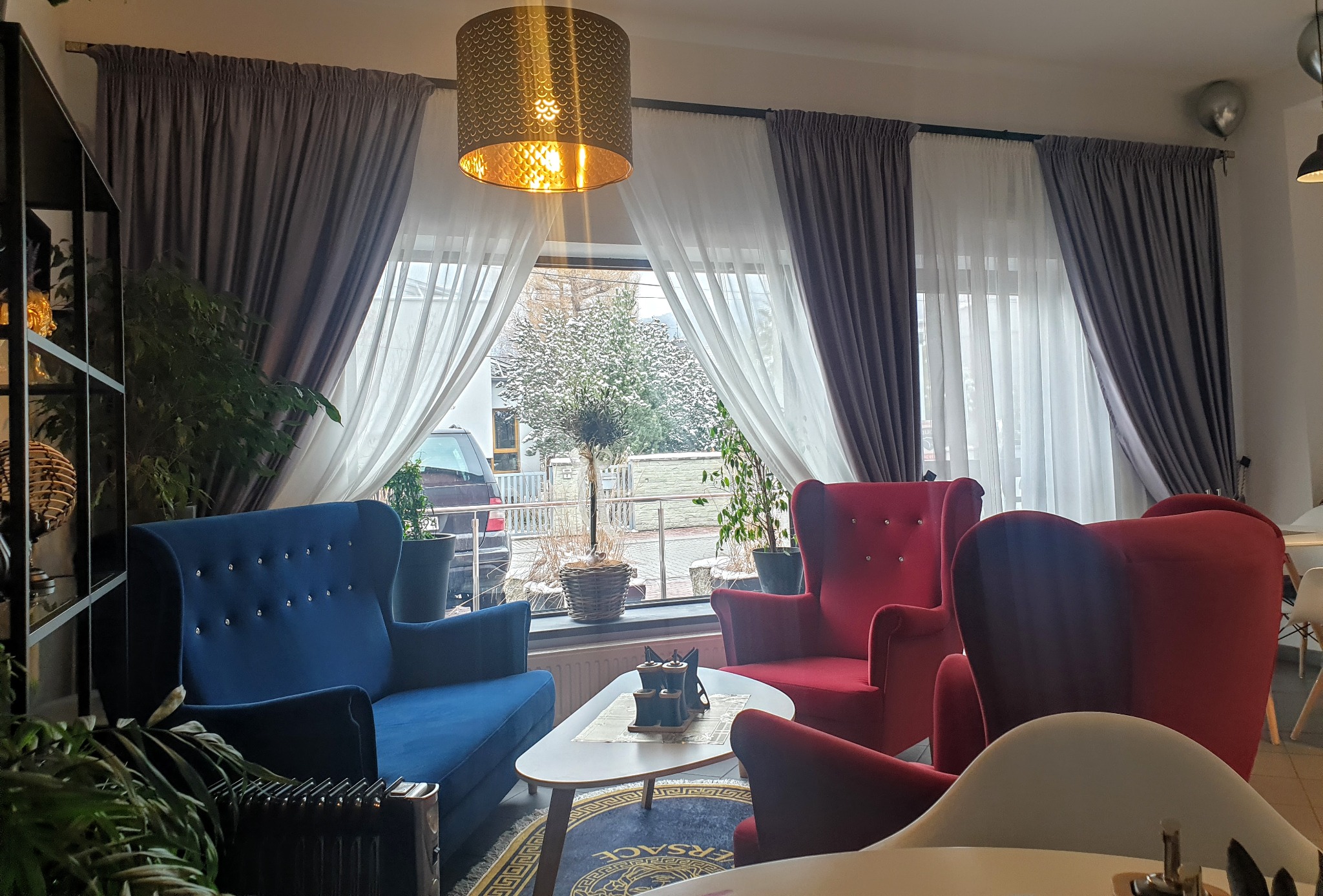 Salon z zasłonami w odcieniach szarości i bieli, fotelami w kolorze czerwonym i niebieskim oraz lampą wiszącą ze złotym kloszem.